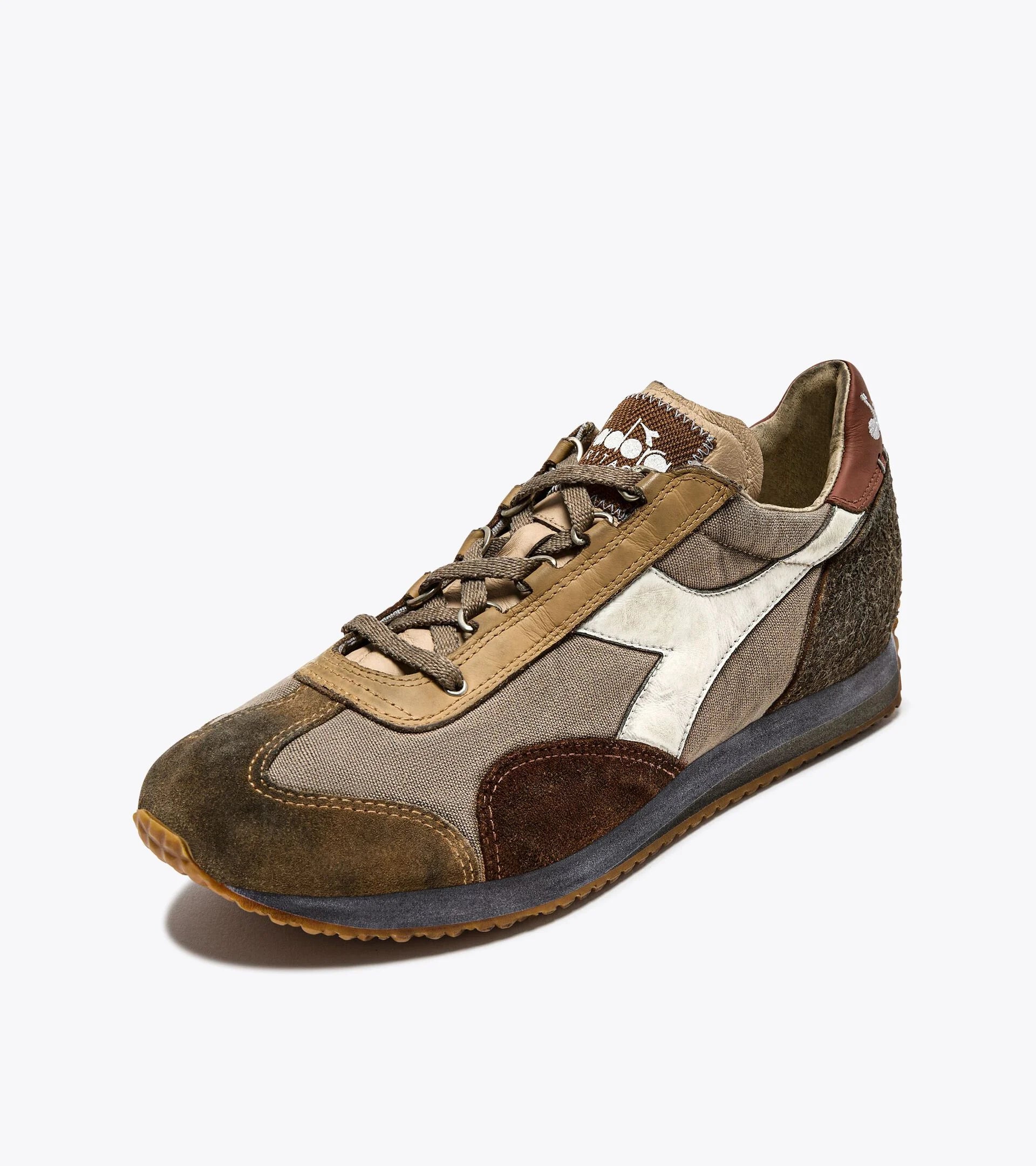 A left-pair upper side view of an Ace Rivington Diadora - Equipe Dirty SW sneaker in Cuban Sand features a mix of brown, tan, and beige suede and fabric upper with a white leather logo stripe on the side. The sneaker has a textured brown heel, a grey midsole, and a gum rubber outsole with a ridged pattern for traction. The laces are brown, and the tongue is made of a similar tan fabric. The heel also has a white logo design for added style.