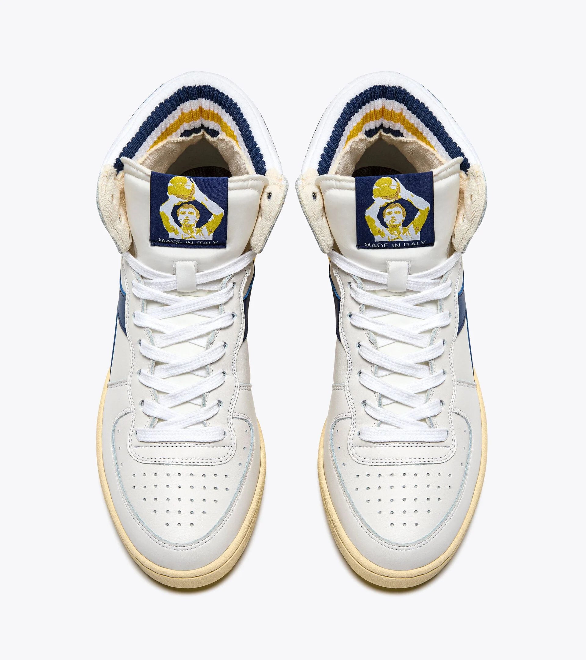 A pair of Ace Rivington Diadora Mi Basket Forty Knit Italia sneakers in white features a high-top design with white laces and perforated toe boxes. The tongue of each shoe has a blue patch with a yellow and white graphic of a person holding a trophy, and the text 'MADE IN ITALY' below the graphic. The inner lining of the shoes has a striped pattern in blue, white, and yellow. The sneakers have a vintage aesthetic with a slightly yellowed sole.