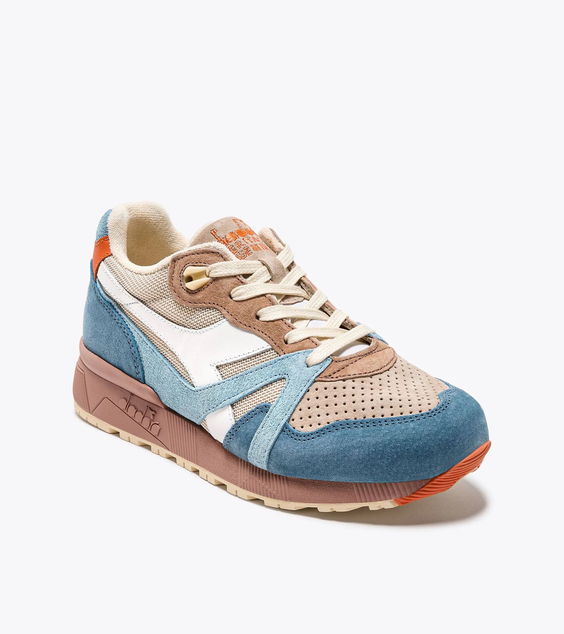 A stylish sneaker from the Ace Rivington Diadora - N9000 Tourney Italia - Blue features a combination of blue, beige, and white suede and mesh materials, with a perforated toe box for breathability. The design includes a white logo on the side, beige laces, and a brown midsole with intricate detailing. The heel has an orange accent, adding a pop of color to the overall design. The sneaker is set against a plain white background, highlighting its unique and fashionable appearance.