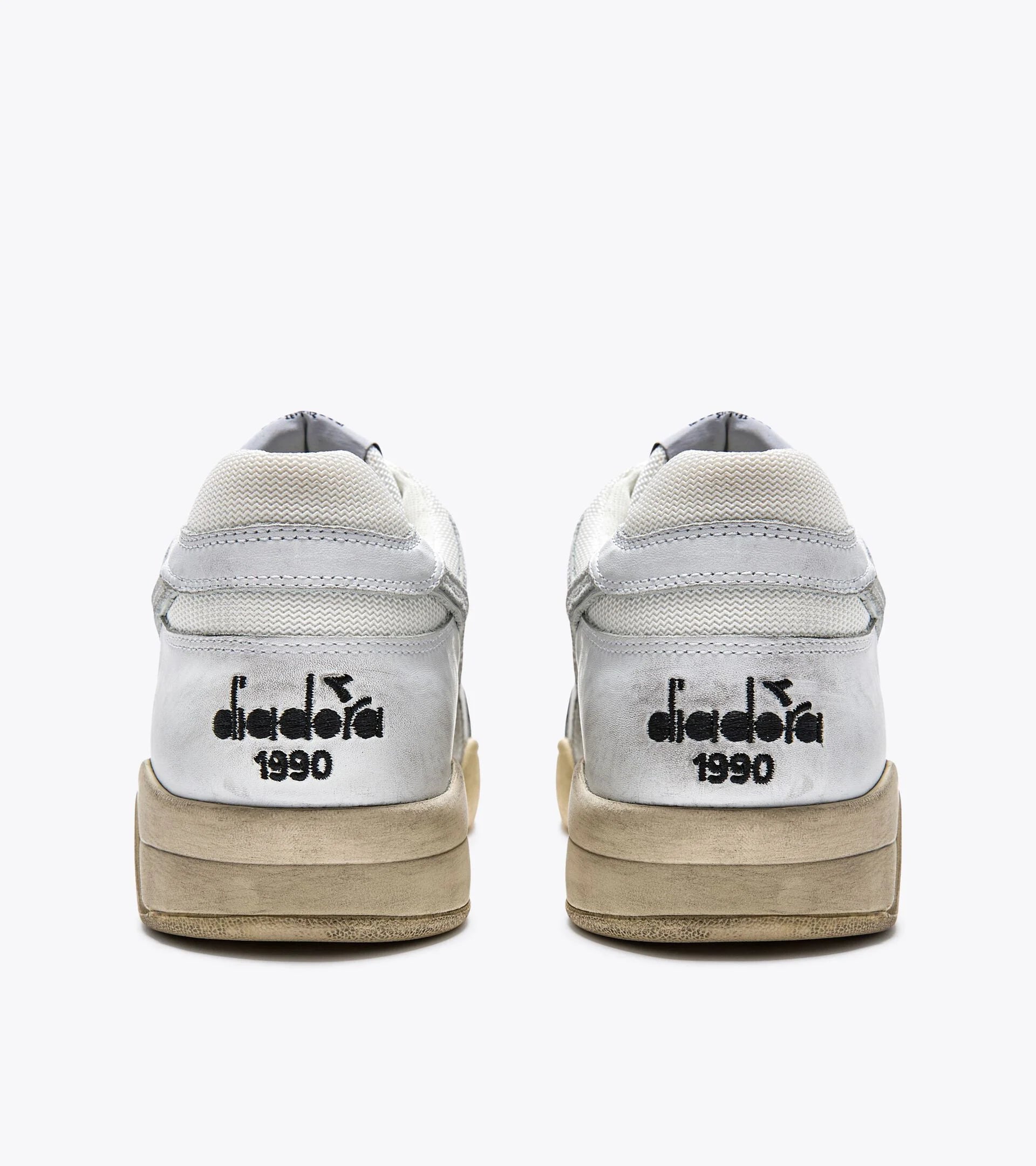 Rear view of a pair of Ace Rivington Diadora B. 560 USED sneakers in white shoes features a worn, vintage look with a slightly dirty sole. The back of each shoe displays the Diadora logo and the year '1990' in black text.