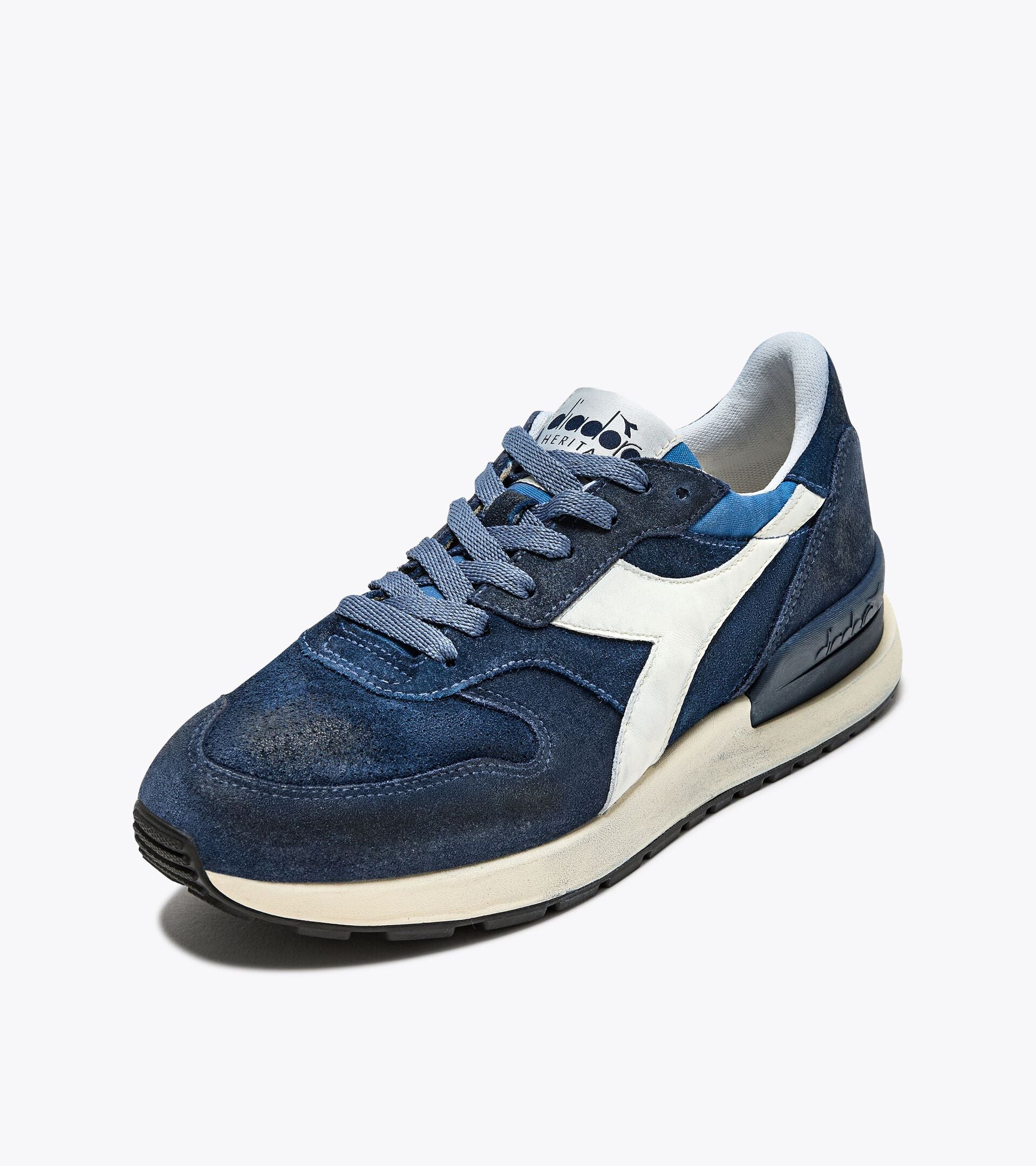 A left-pair front side view of the Ace Rivington Diadora Conquest Suede SW sneakers in Colonel Blue features a dark blue suede upper with a white leather logo stripe on the side, a white midsole, and a black rubber outsole. The laces and tongue are also blue, and the heel has a textured design for added style.