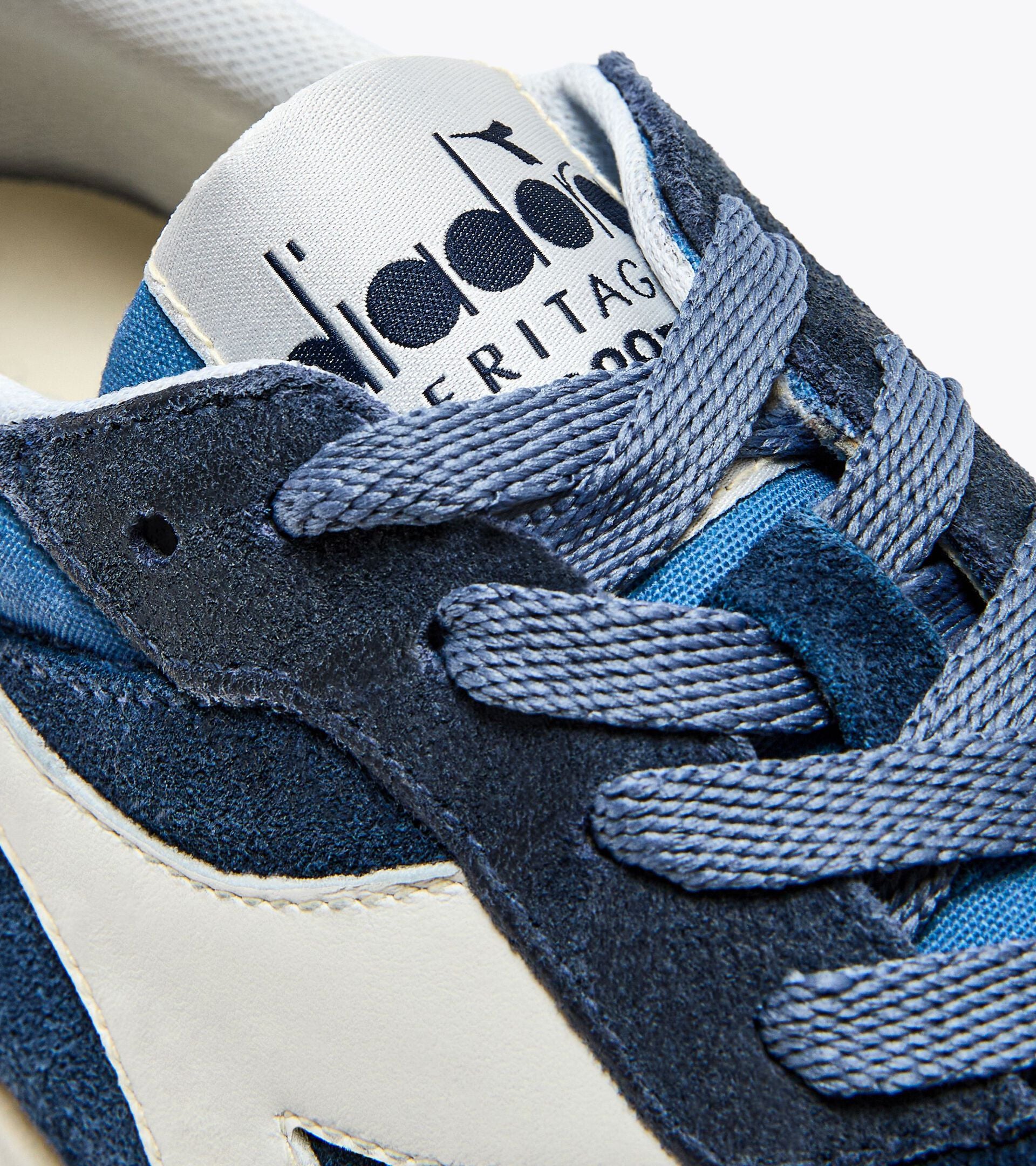 A right-pair close-up of the upper part of an Ace Rivington Diadora Conquest Suede SW sneaker in Colonel Blue highlights the dark blue suede material, blue laces, and the white tongue with the Diadora logo and the word 'HERITAGE' printed on it. The intricate details of the stitching and texture of the suede are visible, showcasing the craftsmanship and design of the sneaker.