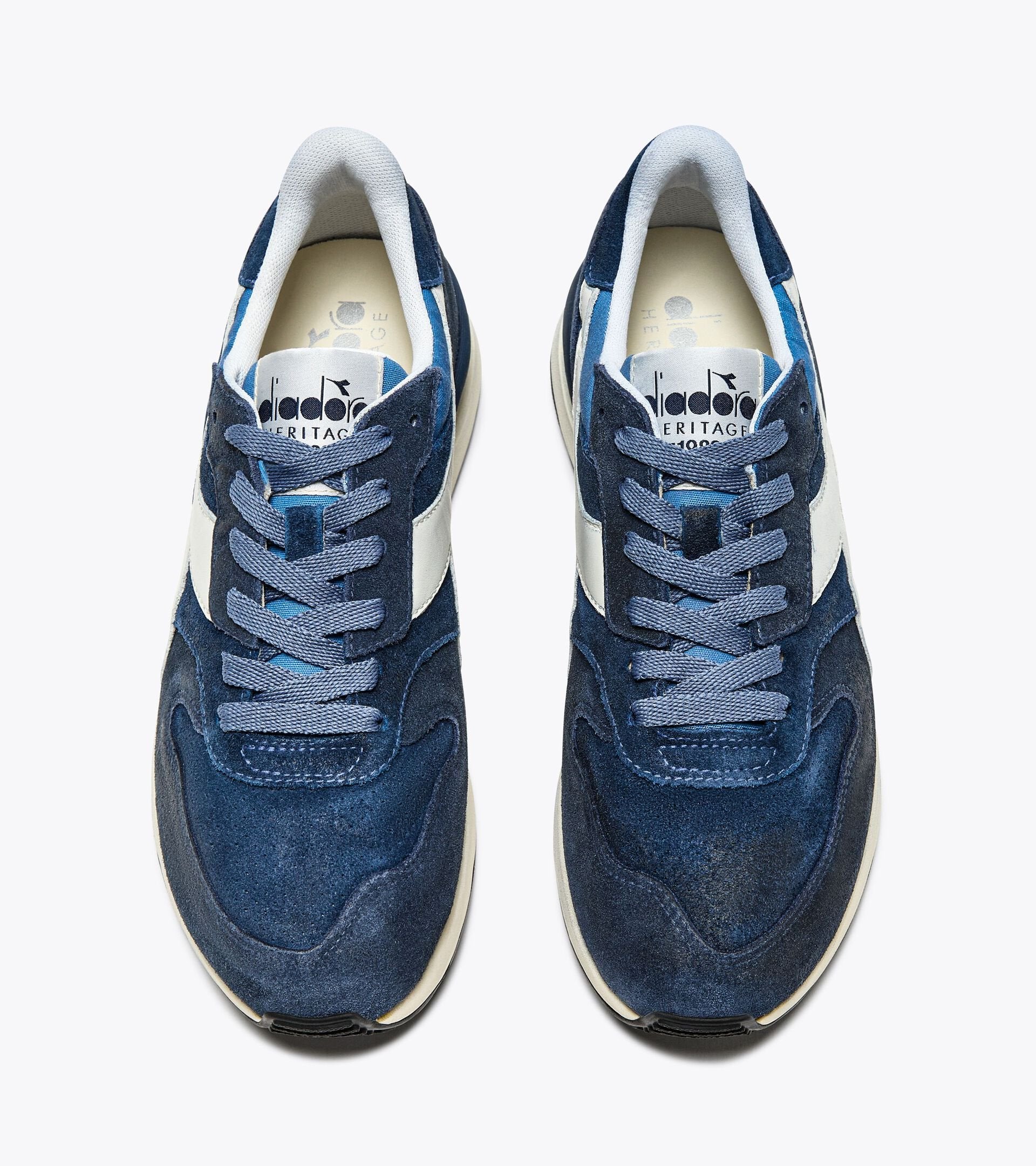 A top view of a pair of Ace Rivington Diadora Conquest Suede SW sneakers in Colonel Blue features a dark blue suede upper with a white leather logo stripe on the side. The laces are blue, and the tongue has the Diadora logo along with the word 'HERITAGE' printed on it. The interior lining is light grey, and the midsole is white with a black rubber outsole.