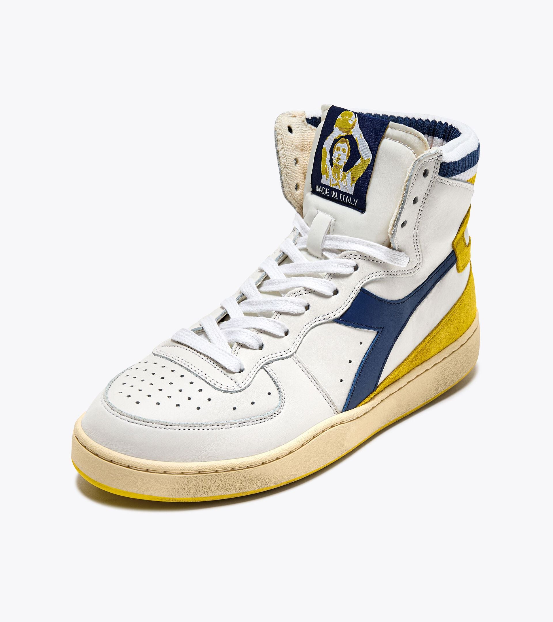 A high-top sneaker from the Ace Rivington Diadora Mi Basket Forty Knit Italia - White shoe features white leather and blue accents, with yellow suede detailing on the heel and sole. The tongue displays a patch with an emblem and the text 'Made in Italy.' The sneaker has white laces and perforations on the toe box for breathability.