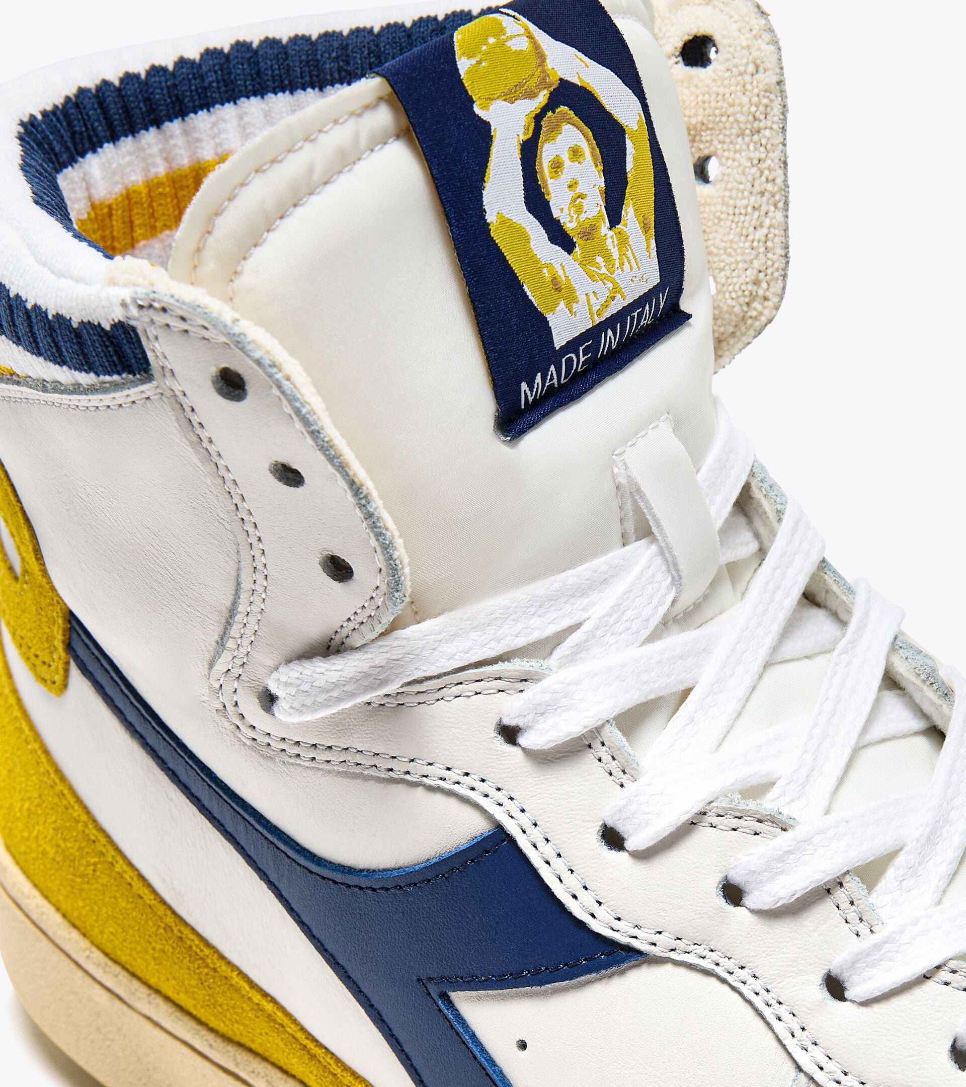 A close-up of the Ace Rivington Diadora Mi Basket Forty Knit Italia sneaker in white shoe features a white leather upper with blue and yellow accents, white laces, and a blue label on the tongue with an illustration of a basketball player and the text 'MADE IN ITALY.'