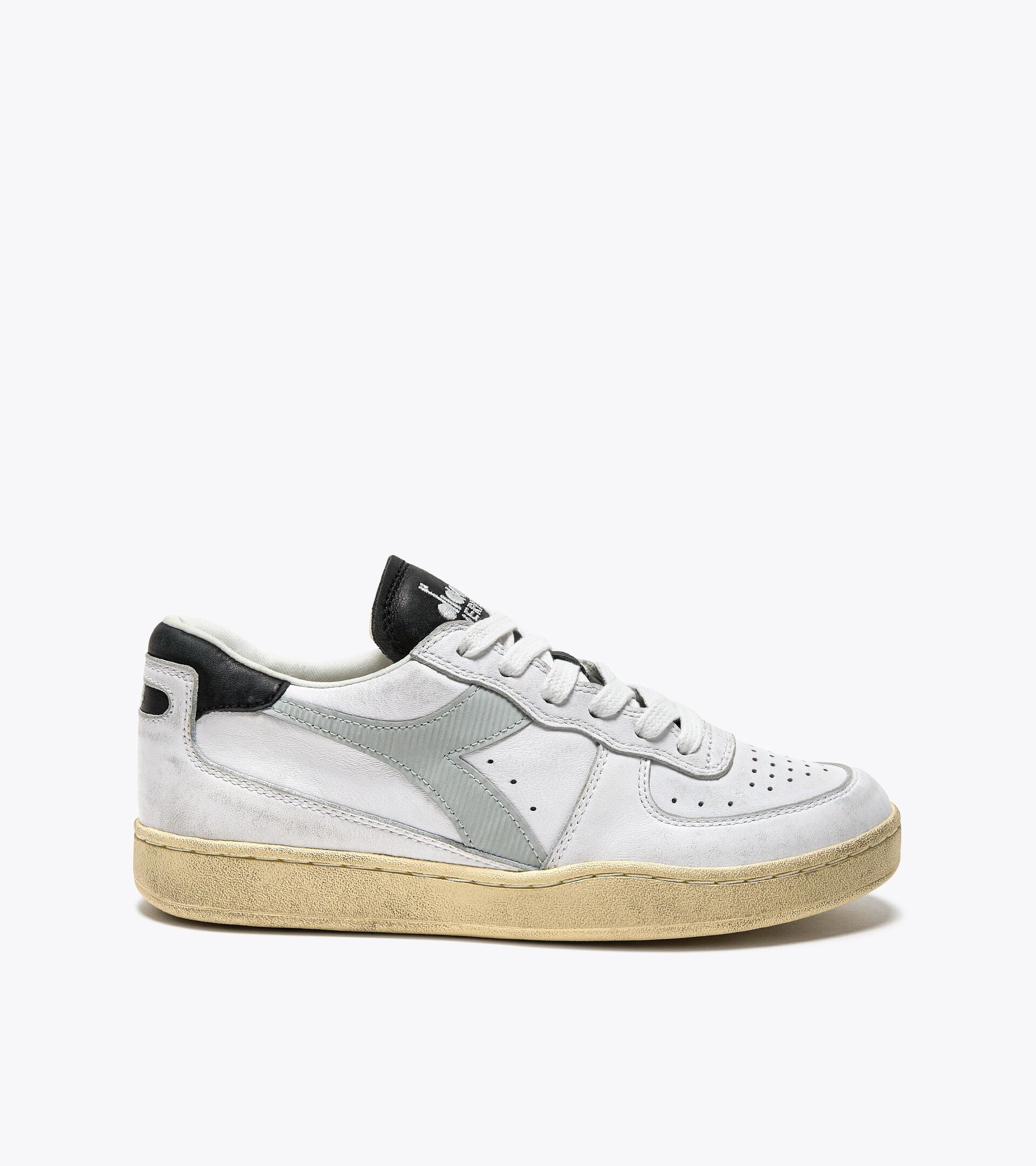 A side view of an Ace Rivington Diadora - Mi Basket Low Used - White/Glacier Gray features a white suede upper with a gray Diadora logo on the side, black accents on the heel and tongue, and a beige, worn-looking sole. The design includes perforations on the toe box and a lace-up closure.