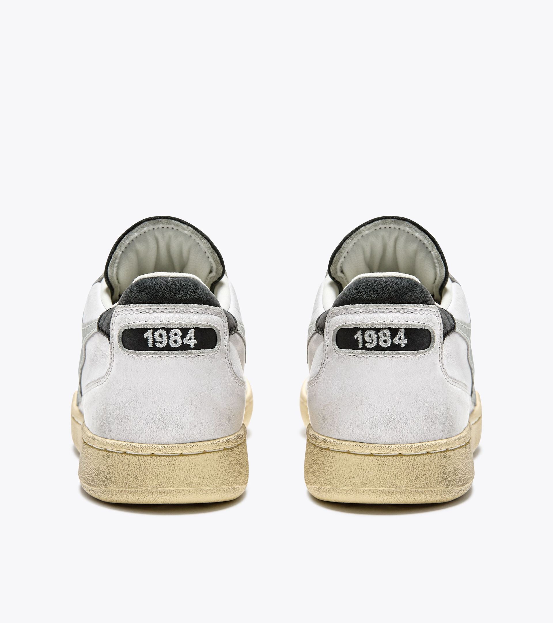 The back view of a pair of Ace Rivington Diadora - Mi Basket Low Used - White/Glacier Gray features a worn look with a beige sole and gray accents. The heel tab displays the year '1984' in white text on a black background, highlighting the vintage inspiration of the model. The shoes also have a suede texture on the heel and a padded collar for added comfort.