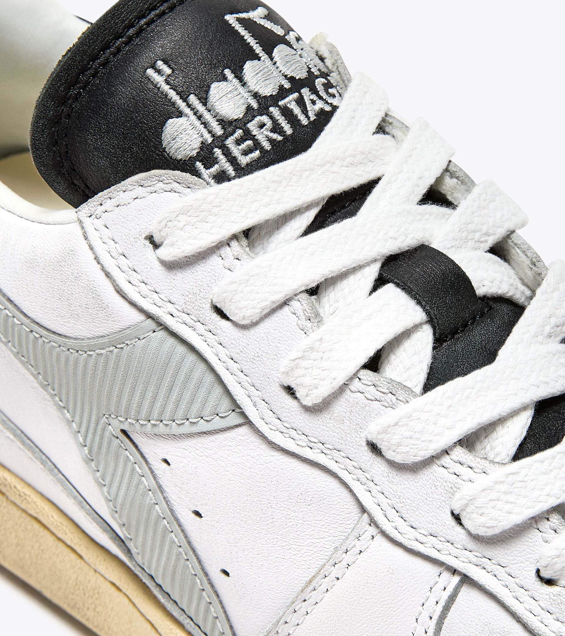 Close-up of the Ace Rivington Diadora - Mi Basket Low Used - White/Glacier Gray highlights the shoe's white leather upper, gray side panel with a ribbed texture and white laces. The black tongue features the Diadora Heritage logo in white. The shoe has a vintage, worn-in look with a slightly yellowed sole.