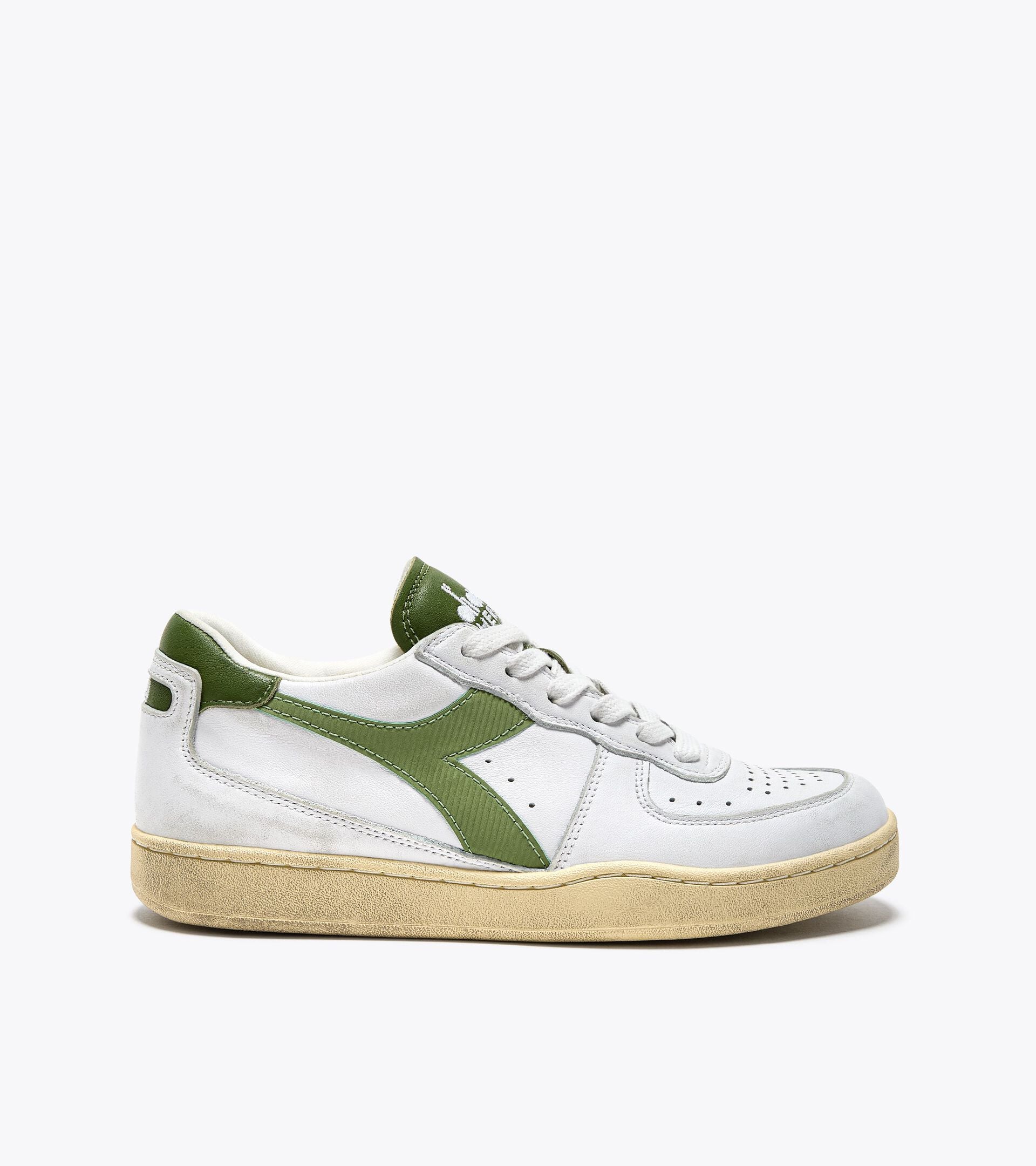 A side view of an Ace Rivington Diadora - Mi Basket Low Used - White/Pesto shoe features a white suede upper with green leather accents, including a green stripe on the side, a green heel tab, and a green tongue with white branding. The sole is beige and has a worn, vintage look. The sneaker has a classic, retro design with perforations on the toe box and side panels for breathability.