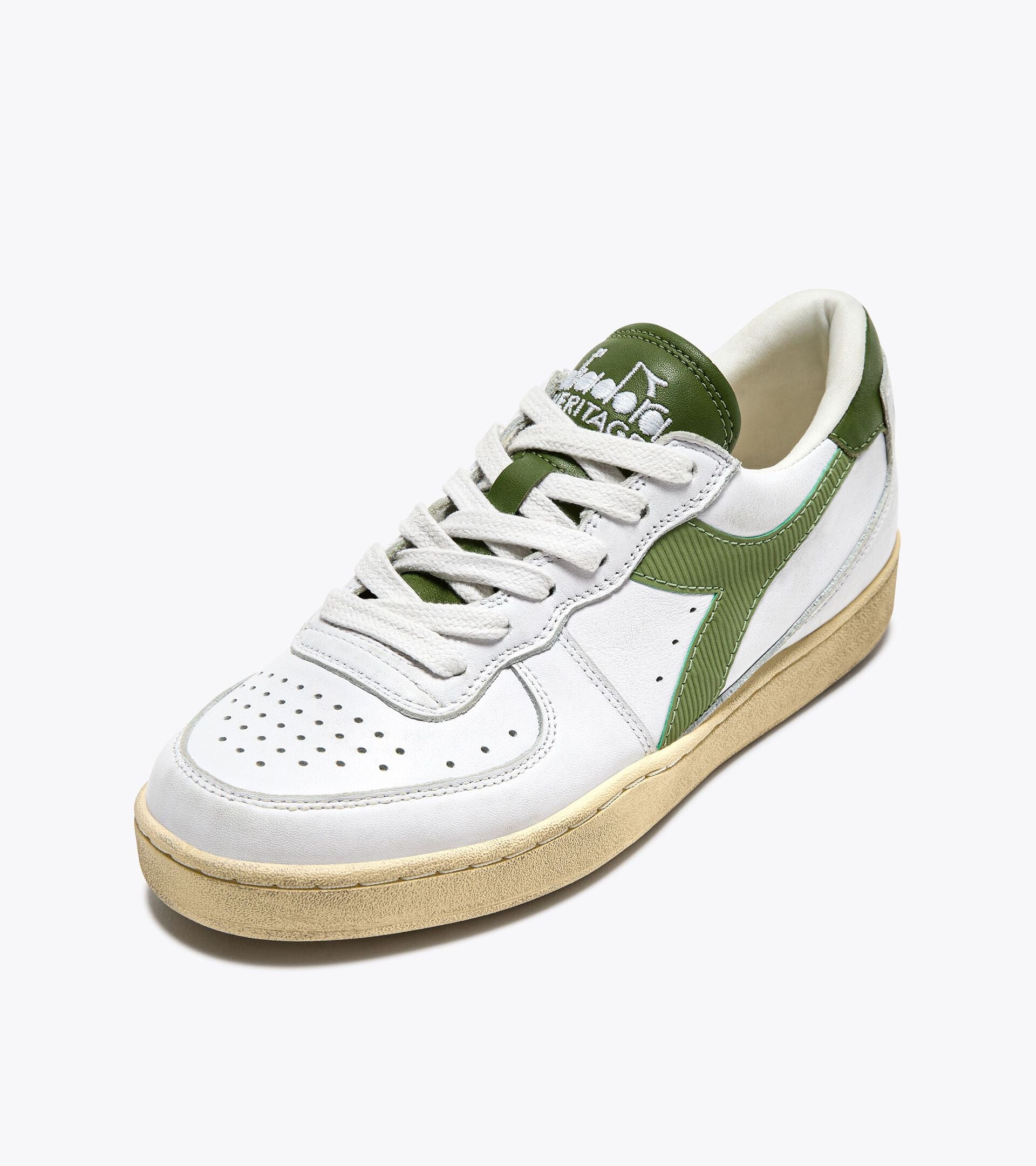 A white and green low-top sneaker from Ace Rivington Diadora - Mi Basket Low Used - White/Pesto shoe features a white leather upper with green accents on the sides, tongue, and heel. The sole is beige with a slightly worn look, and the tongue has the Diadora Heritage logo in white.