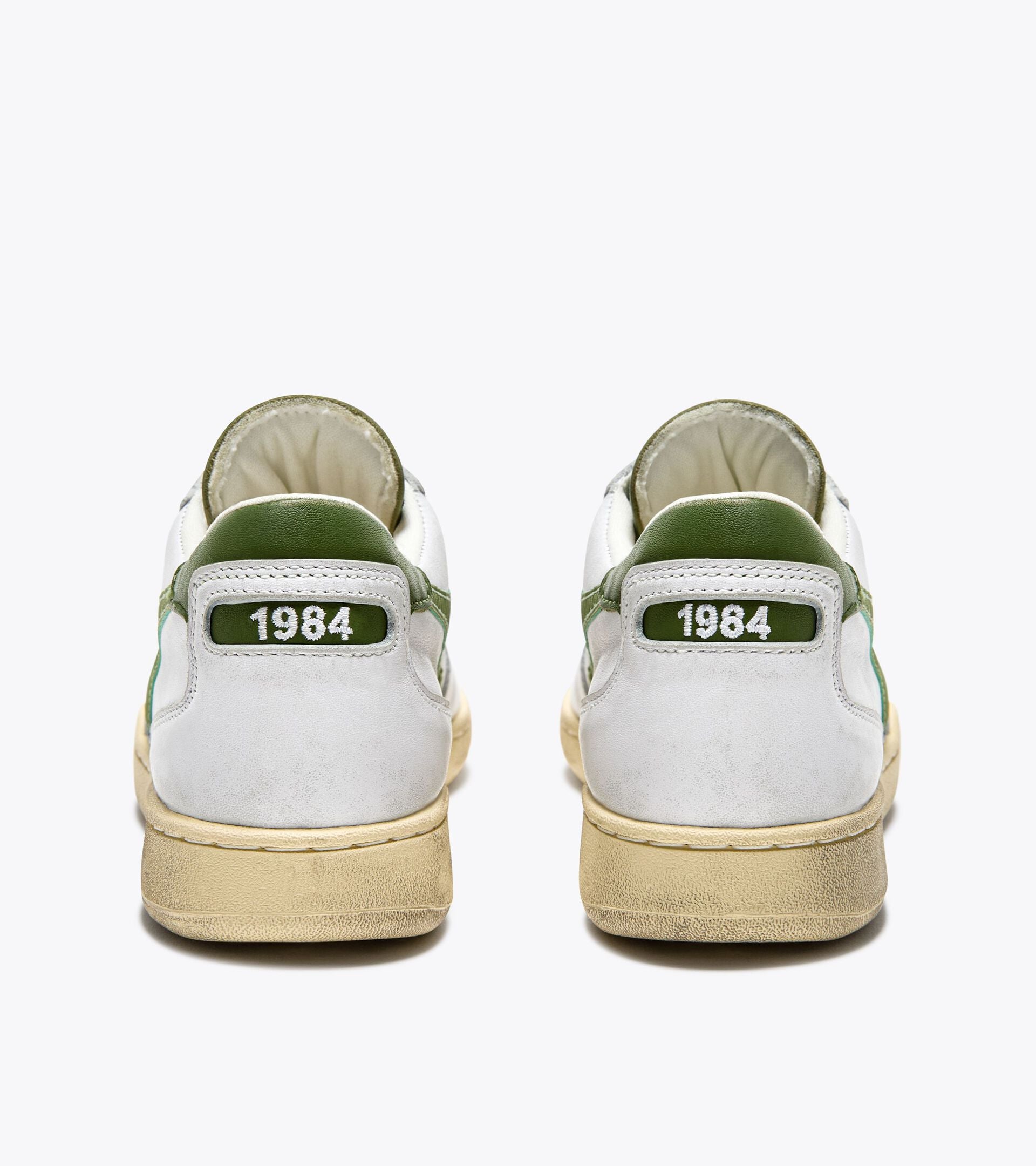 The back view of a pair of Ace Rivington Diadora - Mi Basket Low Used - White/Pesto sneakers have a worn look with a beige sole and green accents. The number '1984' is prominently displayed on the heel tab of each shoe in white text on a green background.