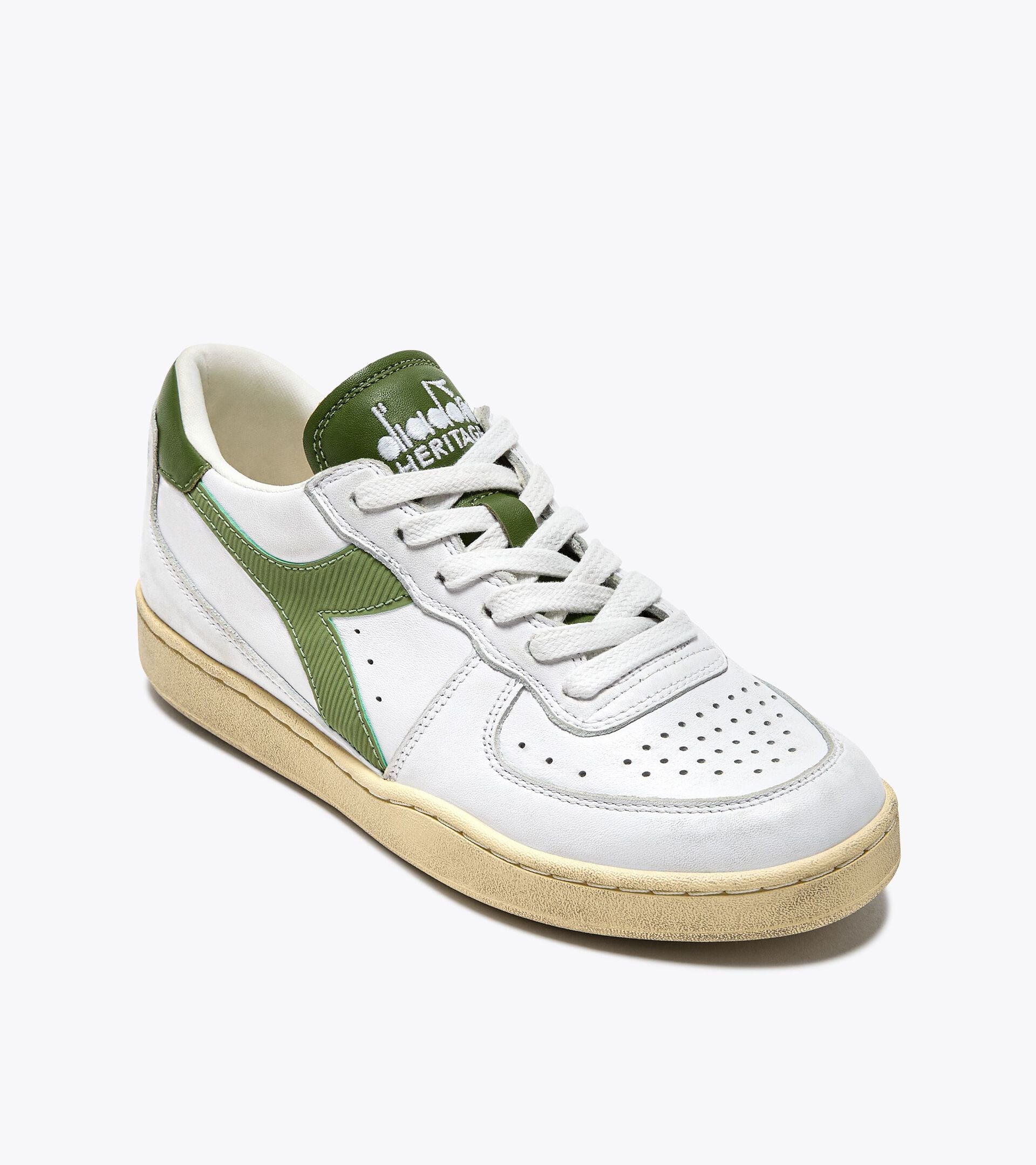 Ace Rivington Diadora - Mi Basket Low Used - White/Pesto shoe angled front view features a white leather upper with green accents, including a green side stripe, green heel tab, and green tongue with the Diadora logo and the word 'HERITAGE' printed on it. The shoe has white laces, a perforated toe box for breathability, and a beige rubber sole that shows signs of wear, giving it a vintage look.