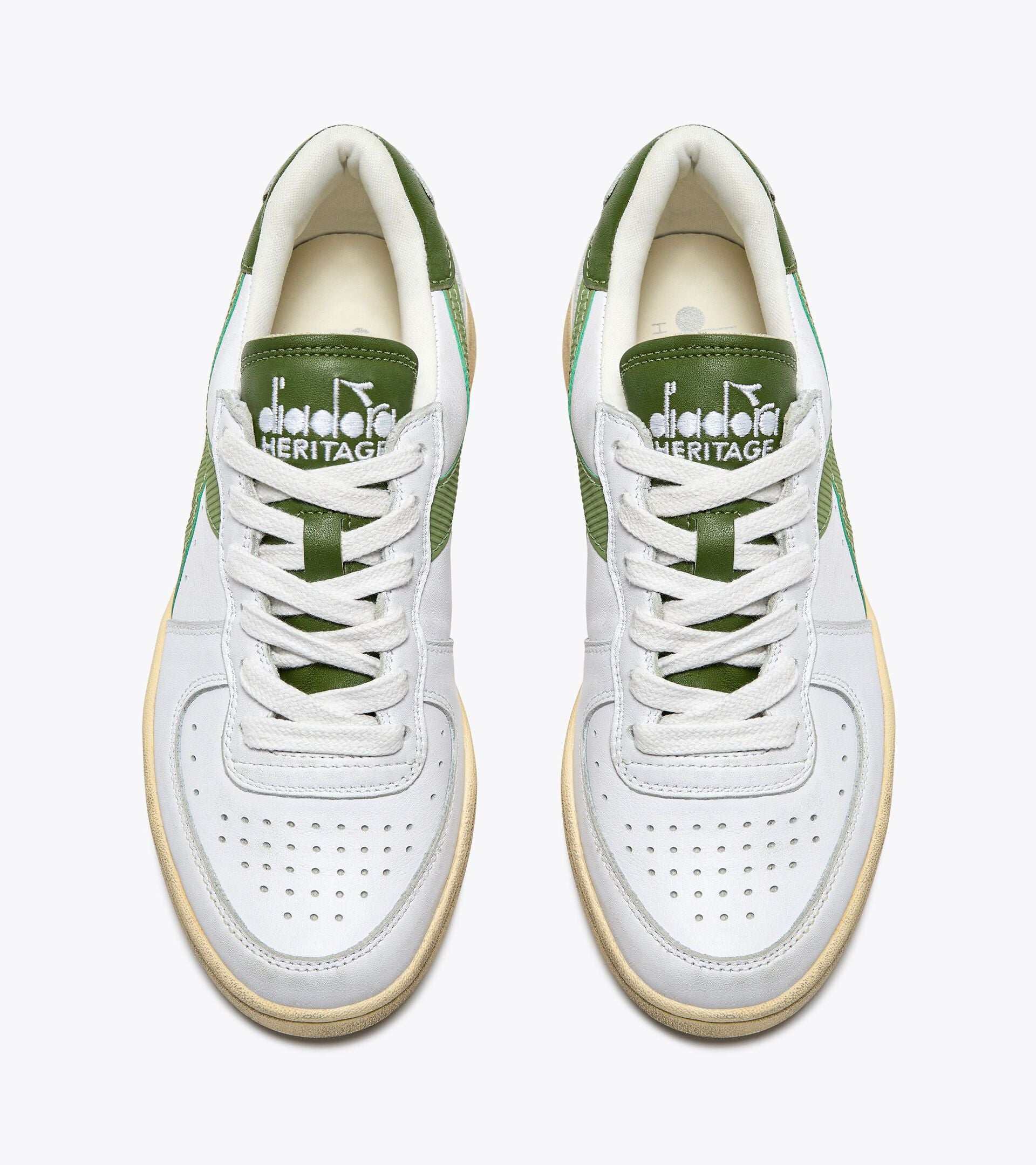 Top view of a pair of Ace Rivington Diadora - Mi Basket Low Used - White/Pesto shoes feature white laces, green tongues with the Diadora Heritage logo, and perforated toe boxes for breathability. The sides have green accents, and the soles have a slightly worn look, adding to the vintage aesthetic.