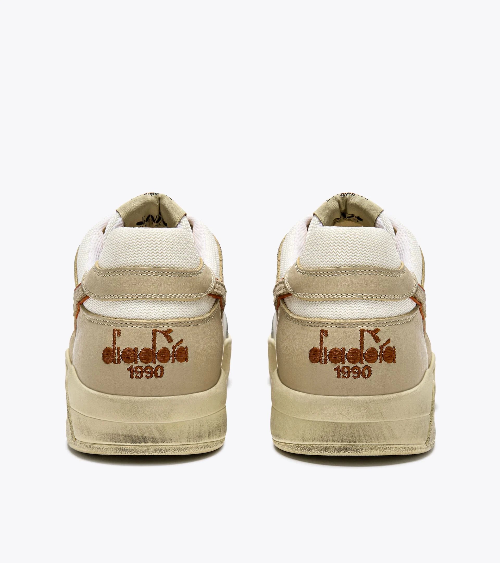 The back view of a pair of Ace Rivington Diadora B. 560 USED - Beige Bleached sneakers feature a beige and white color scheme with visible wear and distressing on the soles and heels. The brand name 'Diadora' and the year '1990' are embroidered in brown on the heel of each shoe. The shoes have a retro design with a padded collar and a textured fabric on the upper part.