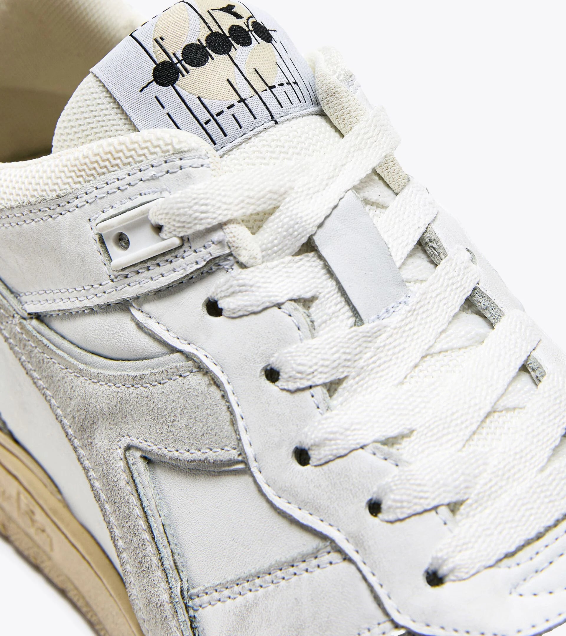 Close-up of a white Ace Rivington Diadora B. 560 USED sneaker, showcasing the detailed stitching, white laces, and a unique graphic design on the tongue. The shoe features a mix of suede and leather materials, with visible wear that adds to its vintage aesthetic.