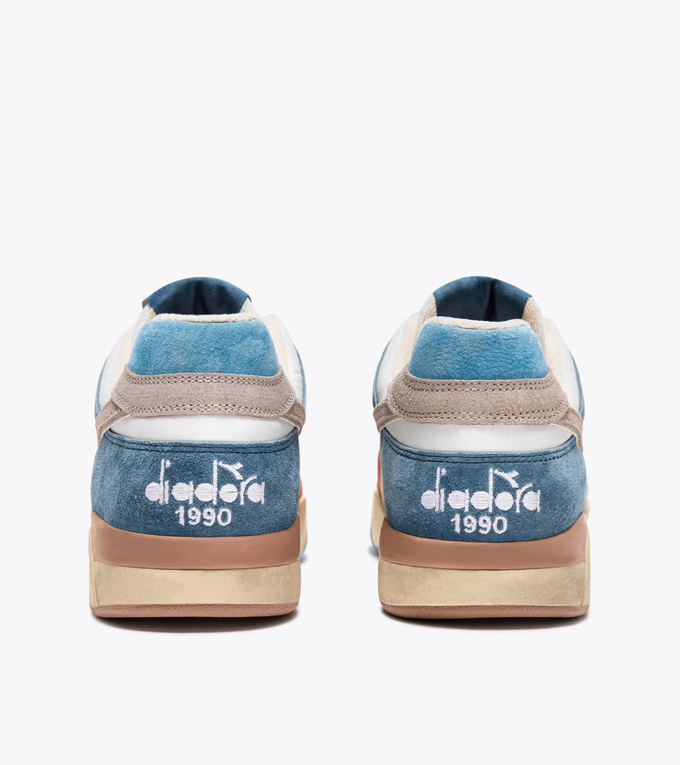 Back view of a pair of Ace Rivington Diadora B. 560 USED - Tourney Italia shoes featuring a combination of blue, beige, and white colors with a suede finish. The heel area has the Diadora logo and the year '1990' embroidered in white. The soles have a slightly worn look, giving the sneakers a vintage or used appearance.