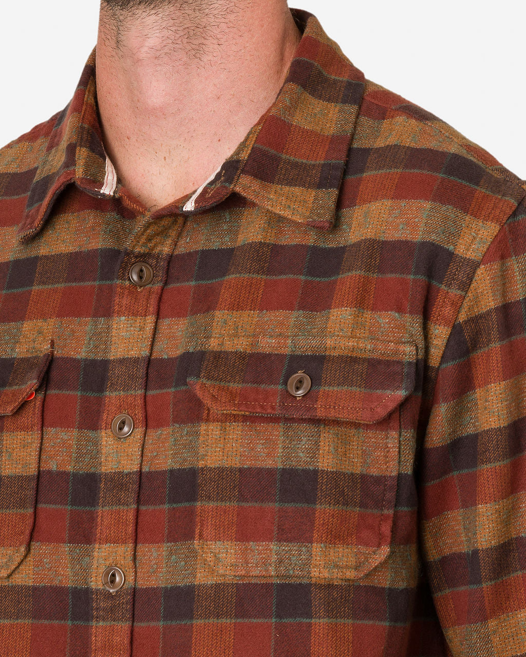 A close-up of a man's neck and chest showcases a plaid clay tile-colored flannel utility shirt featuring a pattern of red, brown, and green squares by Ace Rivington. The shirt includes a collar and buttoned chest pockets.