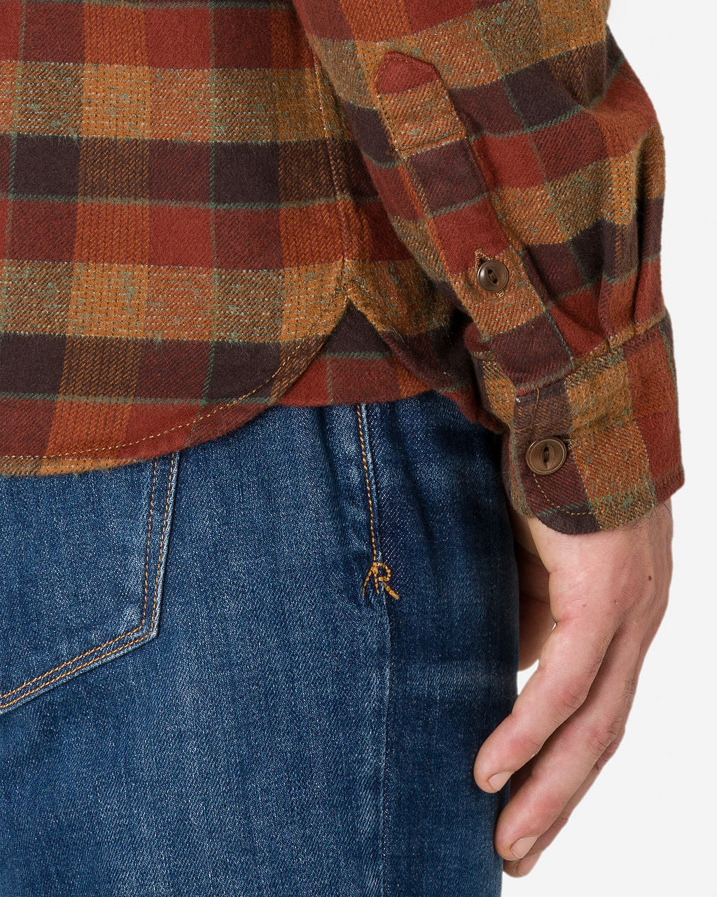 The image presents a close-up view of a man's waistline and the right thigh area, emphasizing a plaid flannel utility shirt in a clay tile color with shades of brown, red, and yellow. The shirt has buttoned cuffs and a curved hem. He is also wearing blue denim jeans by Ace Rivington. The focus of the image is on the details and textures of this casual clothing shirt.