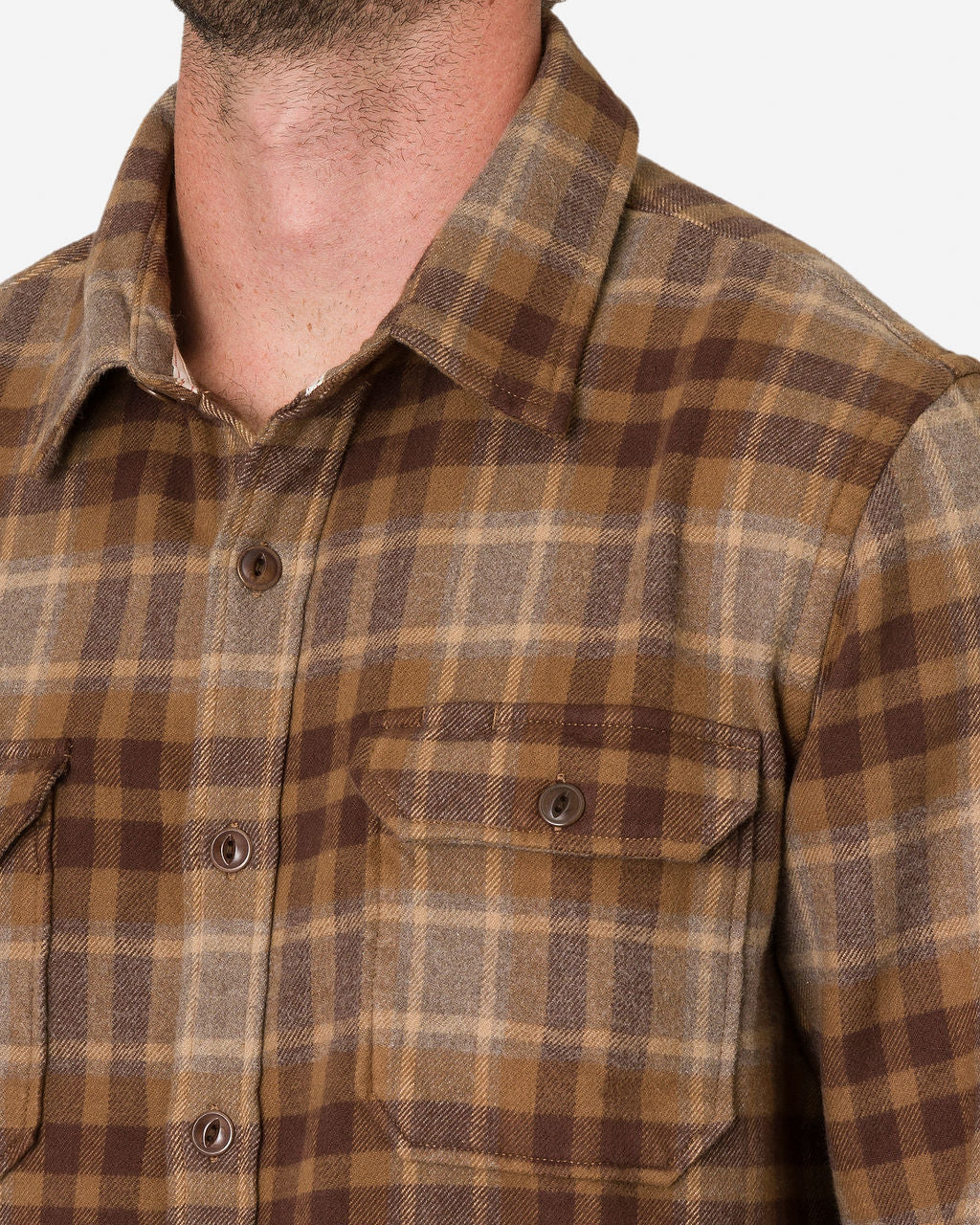 A close-up image of a man showcases his upper torso, highlighting his collar and part of his neck wearing a Desert Bark Flannel Utility Shirt by Ace Rivington, which features a brown and beige plaid pattern. The shirt has a button-up front and two chest pockets with buttoned flaps. The plaid design consists of intersecting lines in varying shades of brown and beige, creating a classic, rustic look.