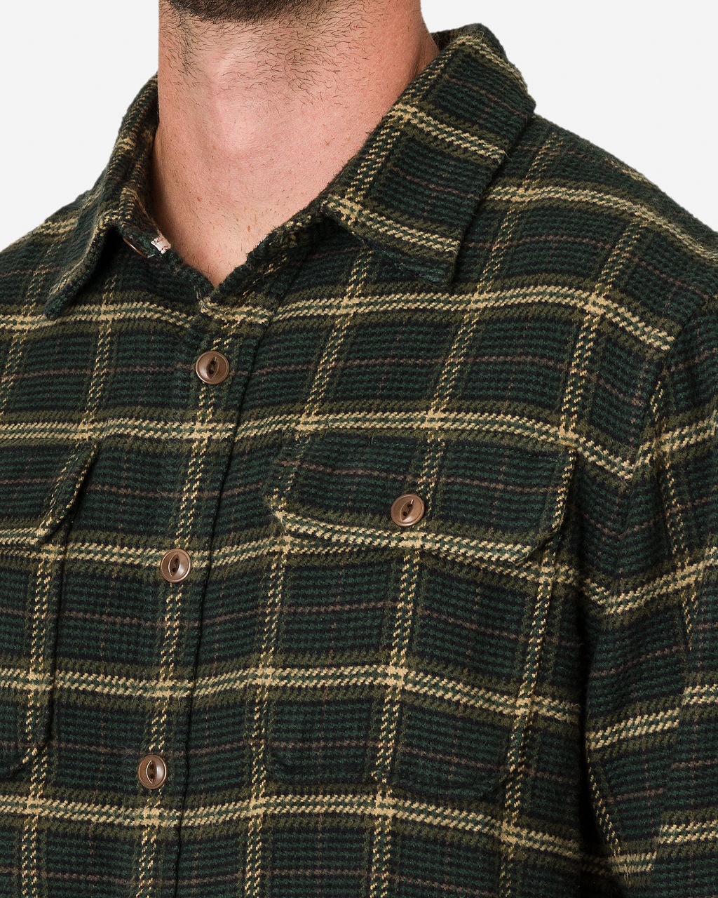 A close-up of a man wearing an Ace Rivington Flannel Utility Shirt - Forest Hound, featuring brown buttons. The shirt includes a collar and a chest pocket with a buttoned flap. The texture and pattern of the fabric are clearly visible, highlighting the design and details on the upper torso of the man.