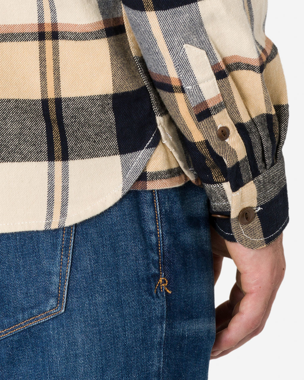 A close-up image of a man wearing a beige, black, gray, and yellow plaid shirt with buttoned cuffs, paired with blue denim jeans. The shirt is untucked, with the person's right hand visible near the side seam of the jeans. 