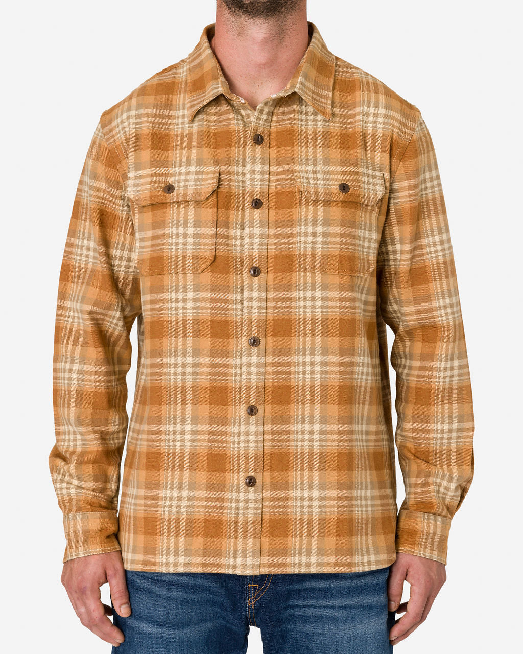 Man wearing a long-sleeved, button-up Ace Rivington flannel utility shirt—sand storm color with a plaid pattern in shades of brown, orange, and beige paired with blue jeans. The shirt has two chest pockets with button flaps. The image focuses on the upper body, showing the shirt's design and pattern details.