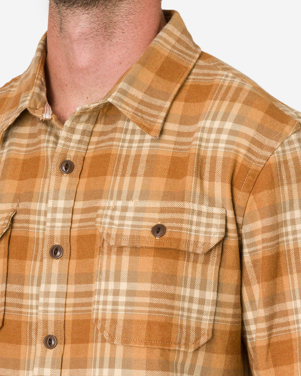 A close-up image of a man wearing a brown, orange, and beige plaid flannel utility shirt—sand storm color by Ace Rivington. Features a button-down front and a chest pocket with a buttoned flap. The pattern consists of intersecting lines forming a checkered design. The image focuses on the upper torso, showing the collar, buttons, and pocket details.