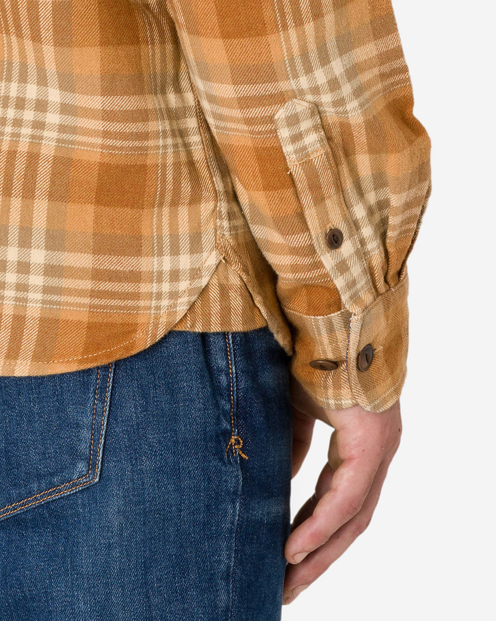 Close-up of a man wearing a flannel utility shirt—sand storm color with a plaid checkered pattern in shades of brown, orange, and beige paired with blue jeans. The sleeve is buttoned at the cuff. 