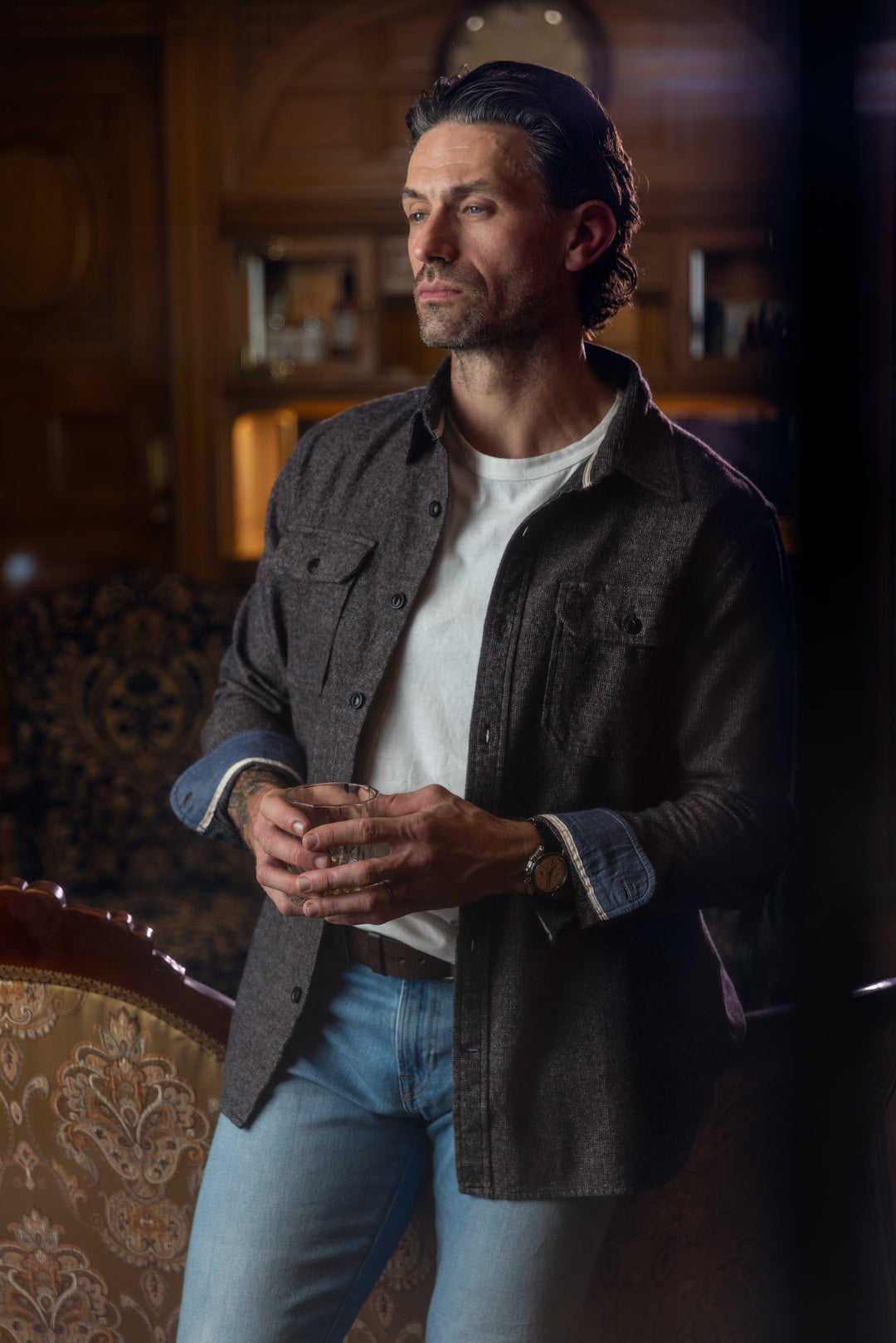 Man standing indoors wearing a white t-shirt under a dark button-up shirt with the sleeves rolled up paired with Ace Rivington - Athletic Taper Denim Jeans - Light Vintage, and a wristwatch. The background features a wooden interior with shelves and decorative elements.