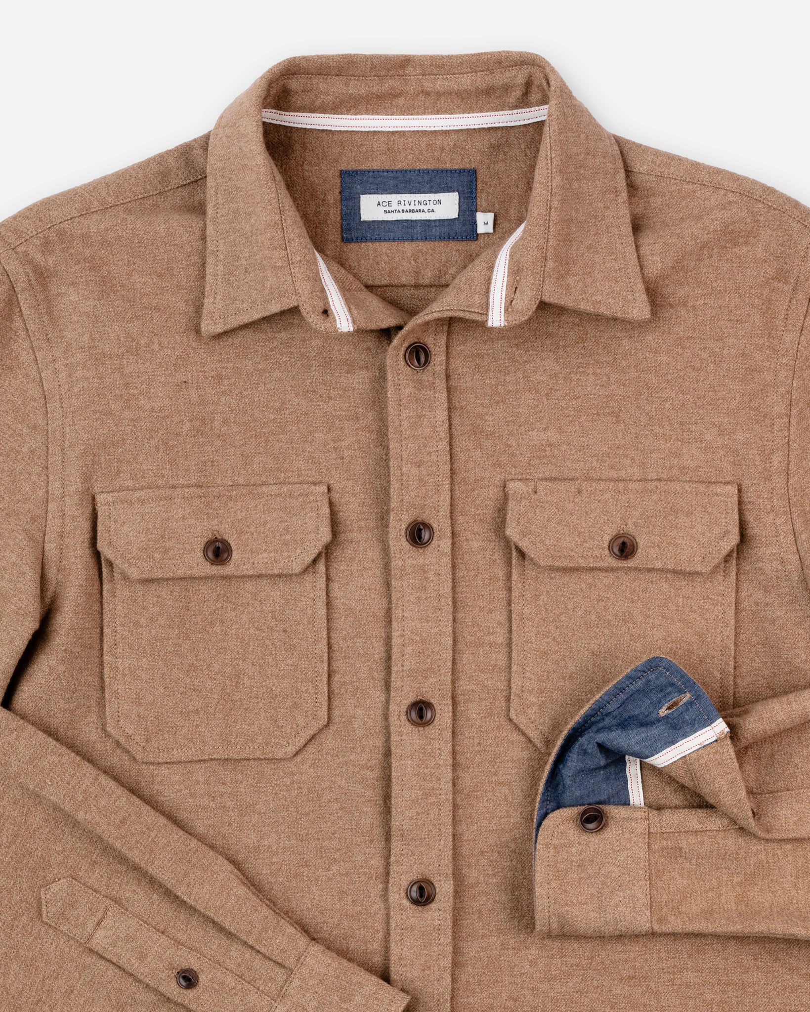 Ace Rivington flannel utility shirt H1 in camel-colored and features two front pockets, each secured with a buttoned flap. It has a collar with white trim on the inside and includes a visible label that reads "Ace Rivington." One sleeve is partially rolled up, revealing a blue inner lining.