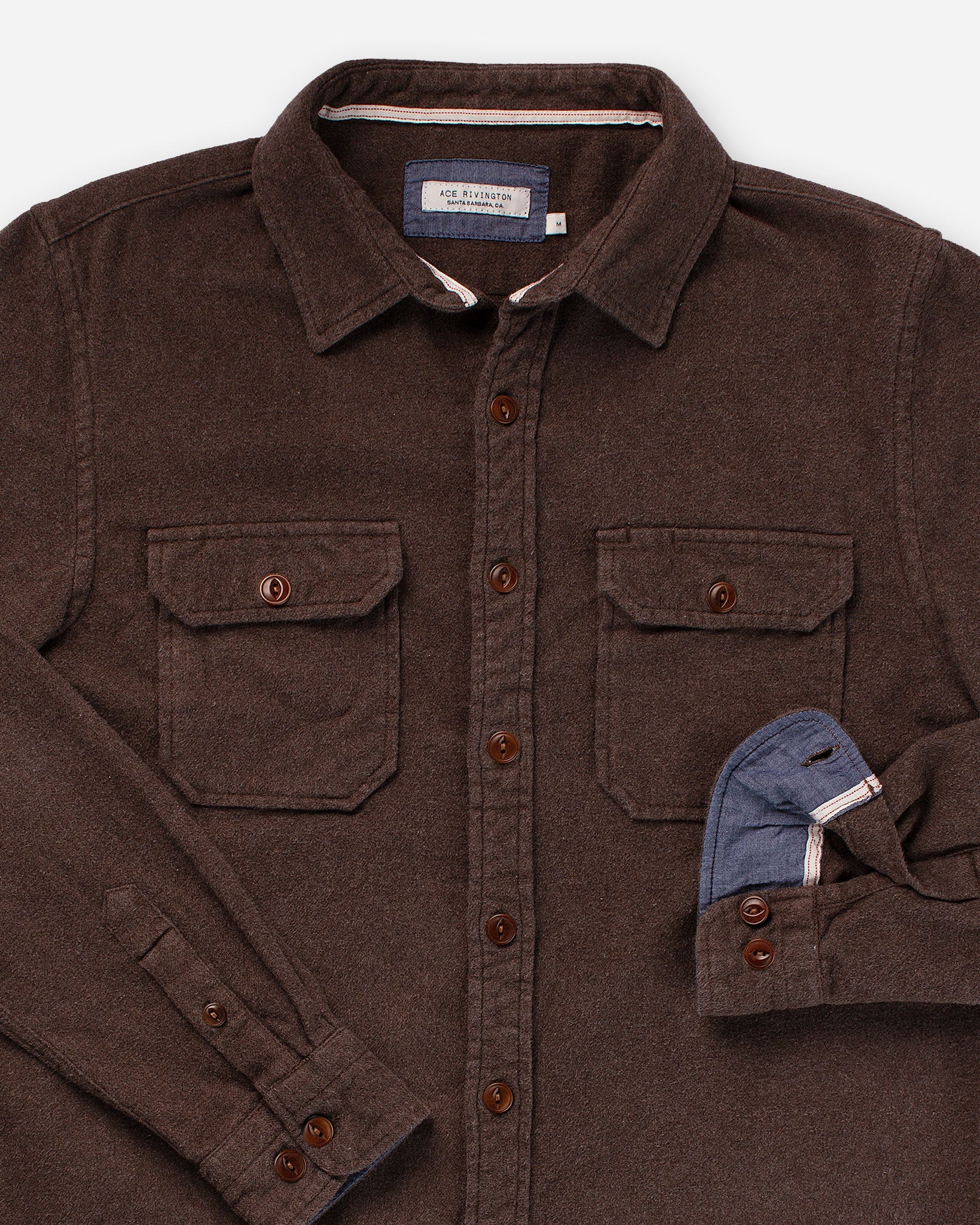 The flannel utility shirt comes in a cedar color and features a button-up design with two chest pockets, each secured by a buttoned flap. It has brown buttons running down the front and on the cuffs. One sleeve is partially rolled up, revealing a blue inner lining. The collar is spread open, and there is a label inside the collar that reads "Ace Rivington."