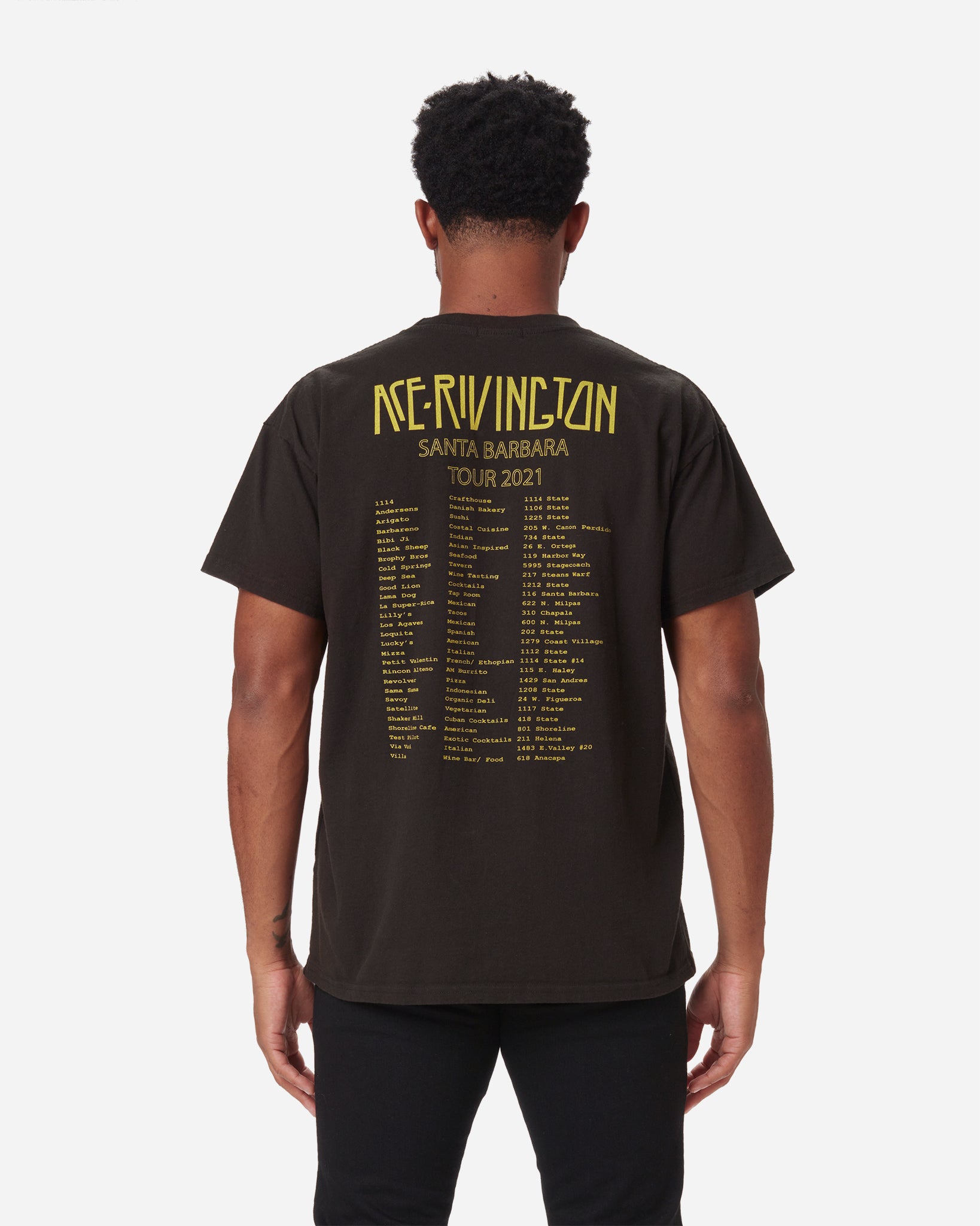 Man shown from the back wearing a black color Ace Rivington- Crewneck SS - 2021 Monster Tour t-shirt features yellow text that reads 'ACE RIVINGTON SANTA BARBARA TOUR 2021' at the top. Below is a list of tour dates and locations printed in yellow, covering most of the back of the shirt. The person is standing with arms relaxed at the sides in the plain white background.