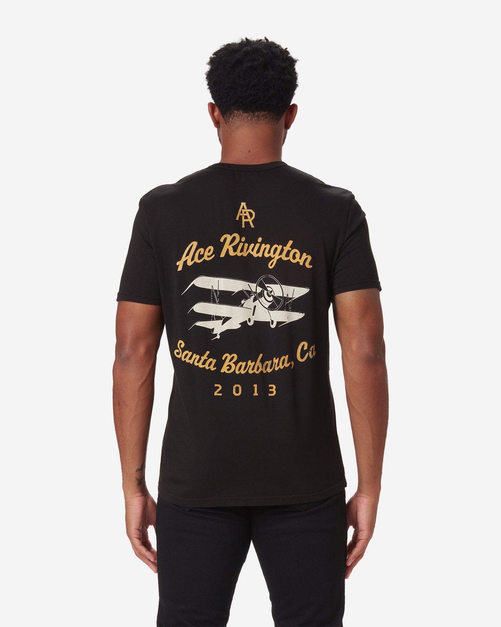 Man wearing a black Ace Rivington Supima Tee Crewneck short sleeves Vintage Flyer t-shirt includes the text 'Ace Rivington' in cursive, 'Santa Barbara, Ca,' '2013,' and a vintage airplane with a pilot. The 'AR' logo is also featured at the top of the design.