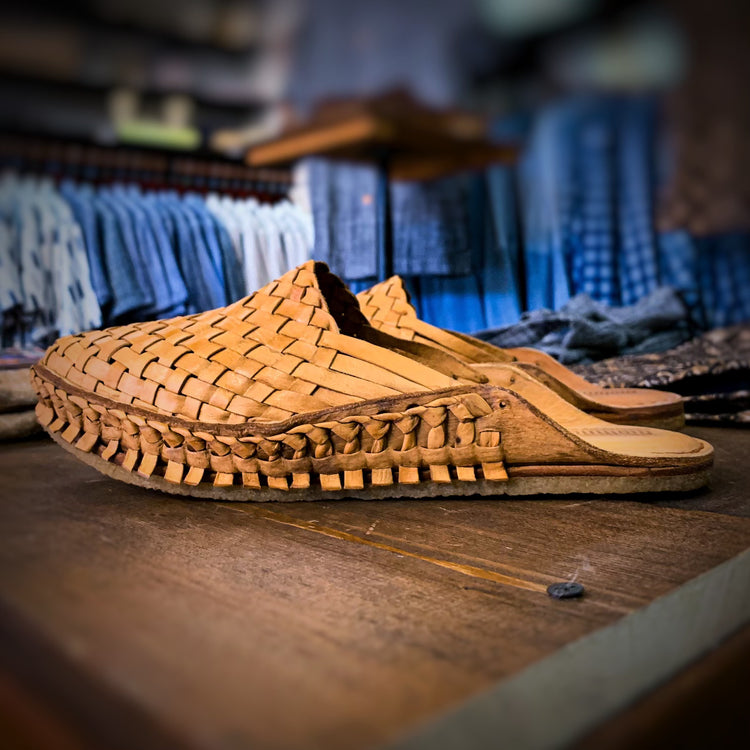 Close-up of Ace Rivington Men's Woven City Slipper in Honey features a detailed woven leather design in a rich honey color. It has a flat sole and an open back, making it easy to slip on. The background shows a blurred clothing store with various garments hanging on racks, adding context to the setting.