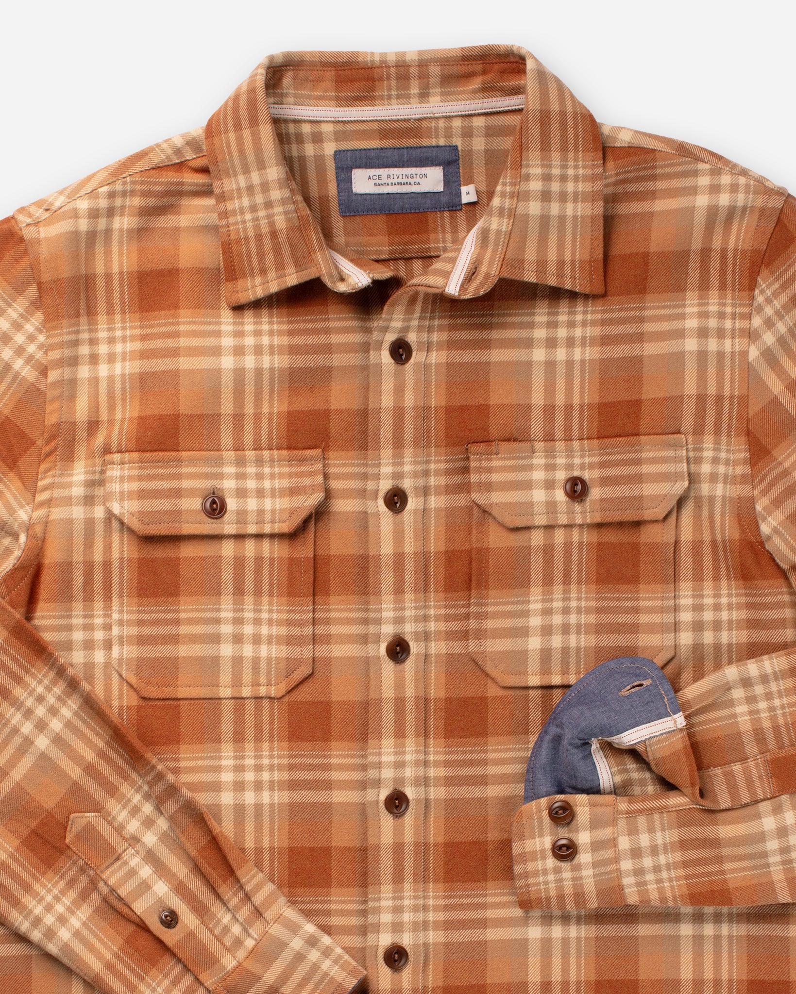 A close-up image of an Ace Rivington long-sleeve, button-up flannel utility shirt—sand storm color with a plaid pattern in shades of brown, orange, and beige with two chest pockets and buttons down the front. The shirt's left sleeve is partially rolled up, revealing a contrasting blue inner lining. The detailed view highlights the shirt's craftsmanship, design elements, and high-quality fabric, making it appealing to potential buyers and fashion enthusiasts.