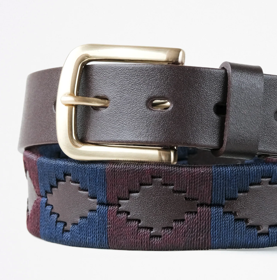 Close-up of the Ace Rivington Macondo Belt - Barolo. The belt features a dark brown leather strap with a gold-colored metal buckle. The woven fabric section displays a geometric pattern in shades of blue and dark-brown, consisting of diamond shapes with a textured appearance.