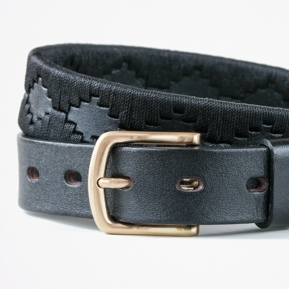 Close-up of the Ace Rivington Macondo Belt - Petrolero. The belt is black with a gold buckle and features an intricate woven pattern in black. The leather strap has visible stitching, highlighting its handcrafted quality. 