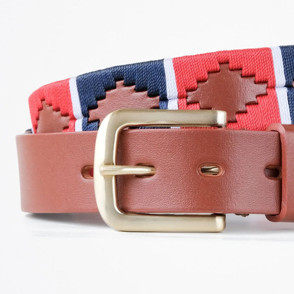 Close-up of the Ace Rivington Macondo Belt - Montanero. This belt features an intricate woven pattern with geometric shapes in red and navy blue, separated by white vertical stripes. It has a brown leather strap with visible stitching and a brass buckle.