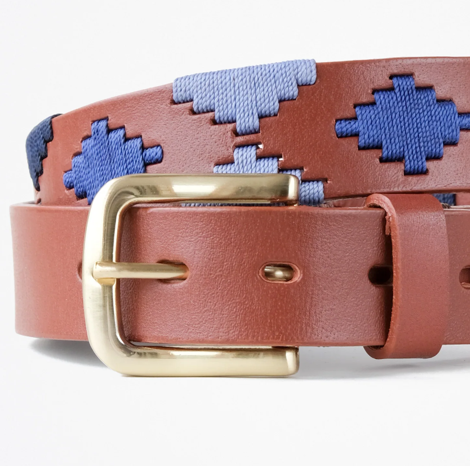 A close-up image of an Ace Rivington Macondo Belts - Almirante. The belt features a brown leather strap with a gold buckle. The leather strap is adorned with a pattern of blue and light blue woven geometric shapes.