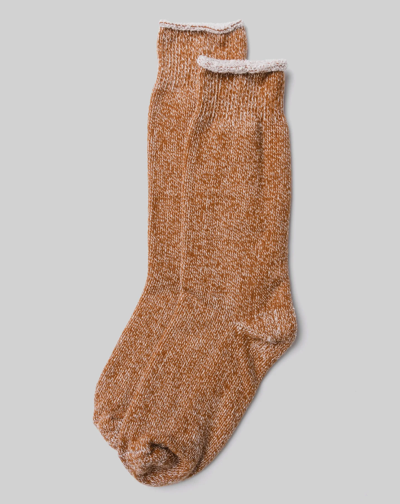 A pair of mustard-colored socks from Ace Rivington The Sunday Sock displayed against a plain background. The socks have a cozy, textured knit with a white trim at the rolled top. One sock is placed slightly on top of the other, showcasing the comfortable and plush design that merges the feel of a sock and a slipper.