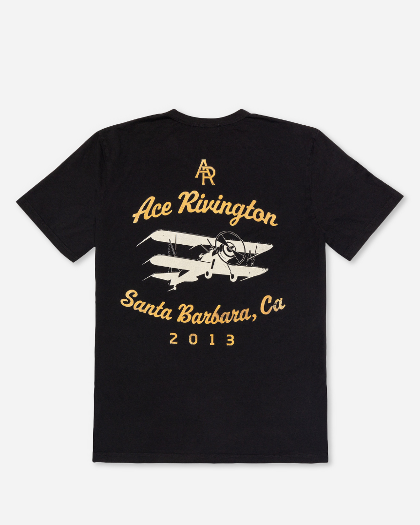 Back view of a black Ace Rivington Supima Tee Crewneck short sleeves - Vintage Flyer t-shirt features a vintage flyer design with a biplane graphic and the text 'Ace Rivington' above the plane, 'Santa Barbara, CA' below, and '2013' at the bottom. The initials 'AR' are printed at the top.