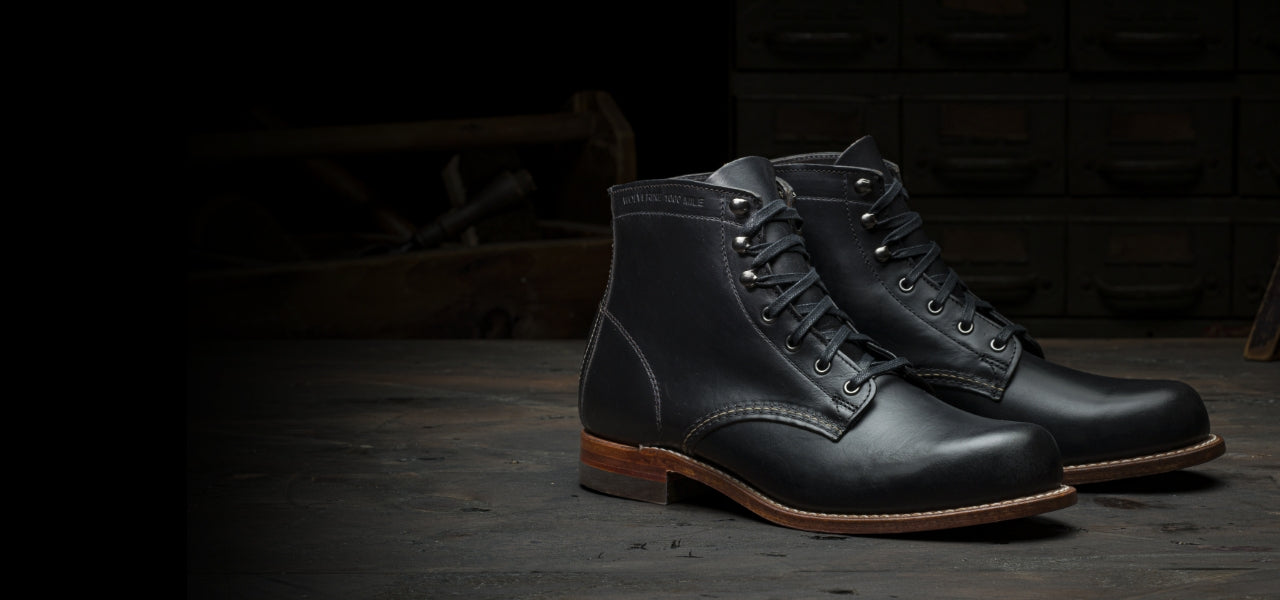 A pair of black leather Ace Rivington Wolverine - 1000 Mile - Plain Toe Original Boot displayed on a wooden surface in a rustic workshop setting. The boots feature brown leather soles, metal eyelets, and visible stitching, highlighting their rugged and classic design.