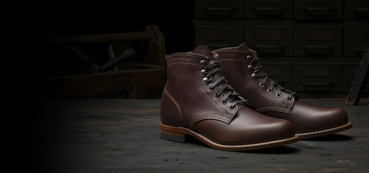 A pair of Ace Rivington Wolverine - 1000 Mile - Plain Toe Original brown leather boots with a classic design, featuring laces and a sturdy sole, placed on a dark wooden surface in a workshop setting. The background includes drawers and tools, highlighting the craftsmanship and artisanal quality of the boots. The well-lit image emphasizes the rich texture and high-quality construction of the leather footwear.