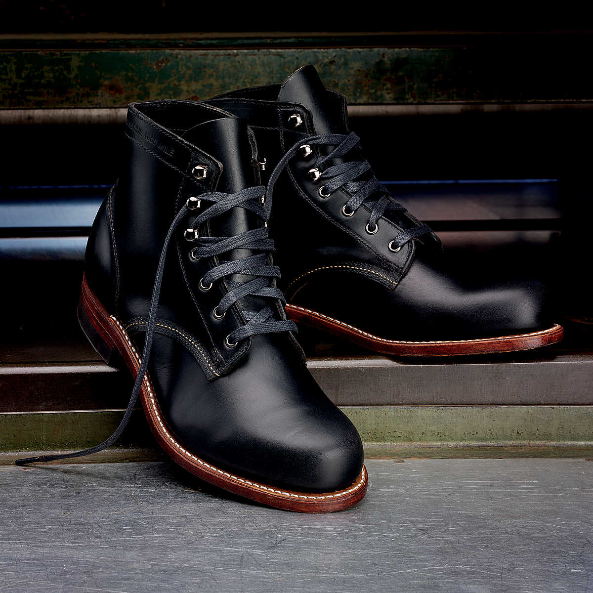 A pair of Ace Rivington Wolverine - 1000 Mile - Plain Toe Original Black Boot with laces and metal eyelets. The boots have a polished finish and feature brown soles with visible stitching around the edges and are displayed on a metallic surface with a greenish background, highlighting their craftsmanship and quality.