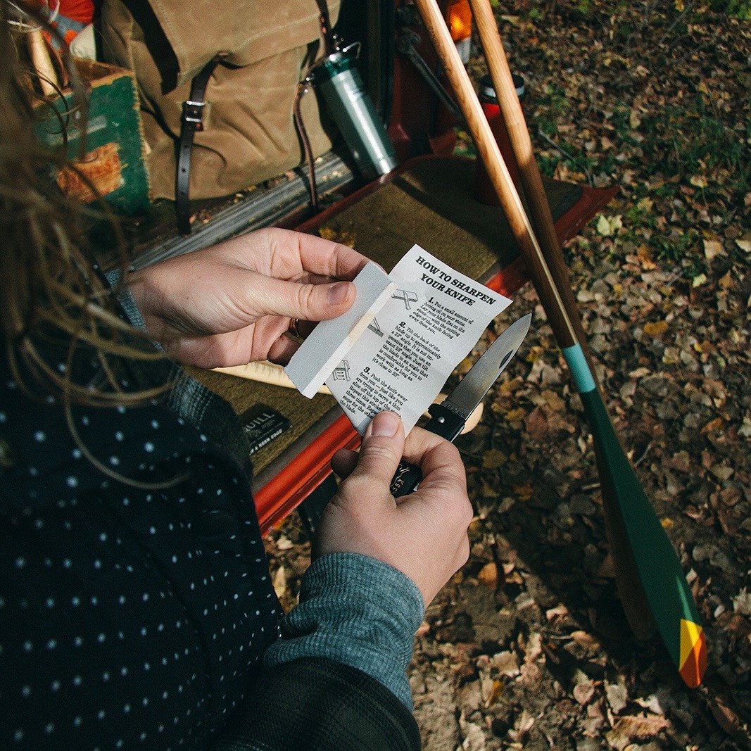 A person outdoors holding a knife and a piece of paper with instructions on how to sharpen a knife. The scene includes fallen leaves and a vehicle with items such as a backpack, a thermos, and paddles. The paper provides step-by-step instructions for sharpening a knife, emphasizing the practical use of the Ironquill Hook and Knife Sharpening Stone.