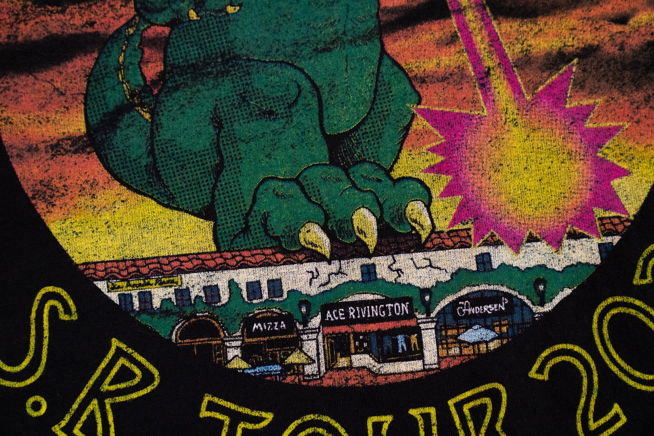 Graphic design on a black Ace Rivington Heavy Tee - Crewneck SS from the 2021 Monster Tour t-shirt. The design features a large green monster's foot claw gripping a row of buildings, including a storefront labeled 'Ace Rivington.' The background shows a dramatic sky with a bright, starburst-like explosion. The text 'S.R Tour 2021' is partially visible at the bottom of the image.