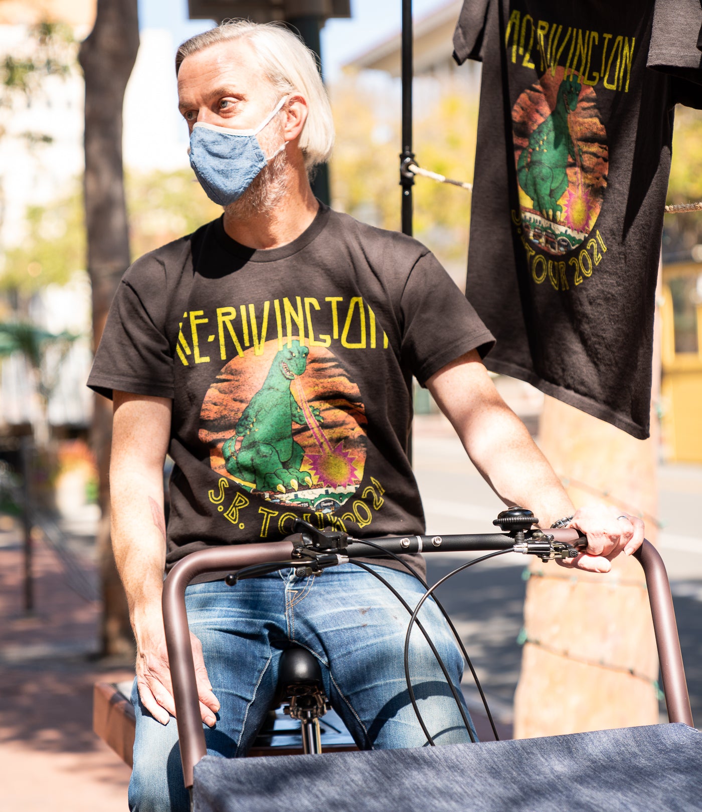 Man sitting on a bicycle, wearing a black t-shirt with a colorful graphic design featuring a green Godzilla-like dinosaur and the text 'ACE RIVINGTON S.B. TOUR 2021.' Another identical t-shirt is hanging in the background. The scene is set outdoors on a sunny day, with blurred trees and buildings in the background. The image highlights the Ace Rivington Heavy Tee - Crewneck SS from the 2021 Monster Tour collection in black color.