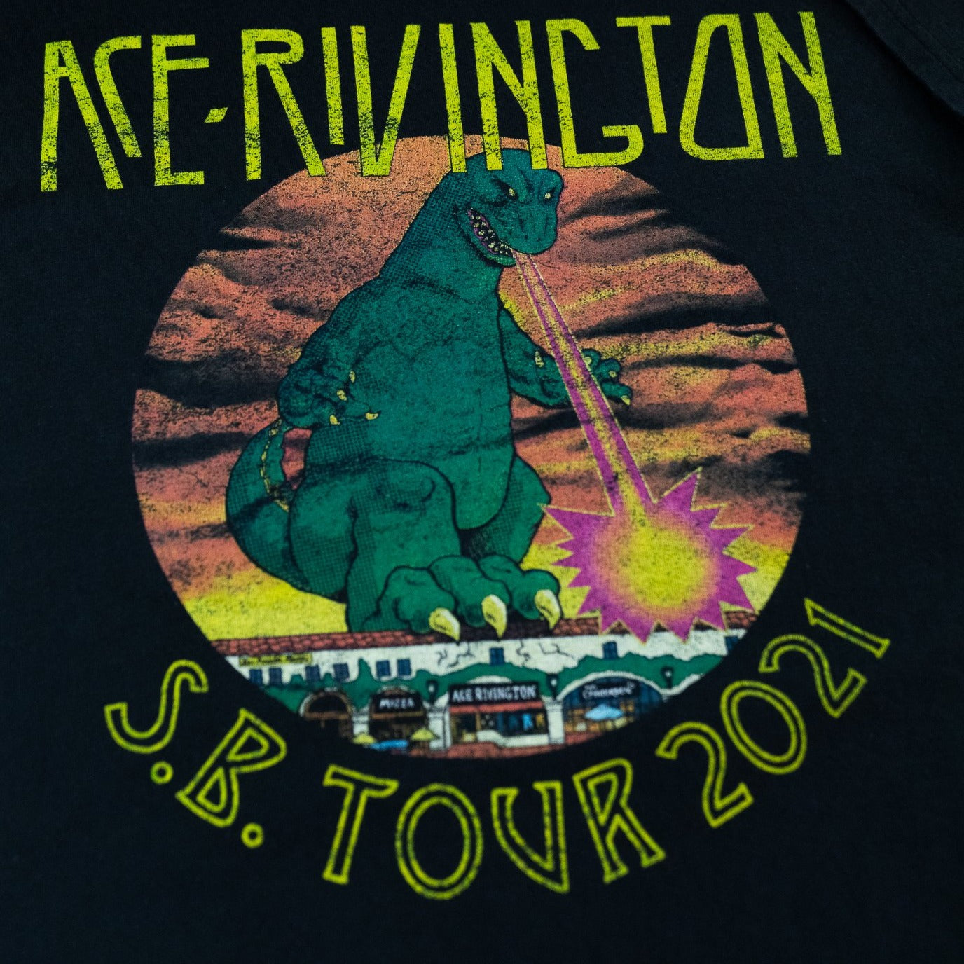 Ace Rivington Heavy Tee - Crewneck SS - 2021 Monster Tour Black color t-shirt. The design features a large green monster, reminiscent of Godzilla, shooting a laser beam from its mouth toward a cityscape. The background includes a colorful sunset with clouds. The text 'ACE RIVINGTON' is displayed at the top in bold, stylized yellow letters, and 'S.B. TOUR 2021' is written at the bottom.