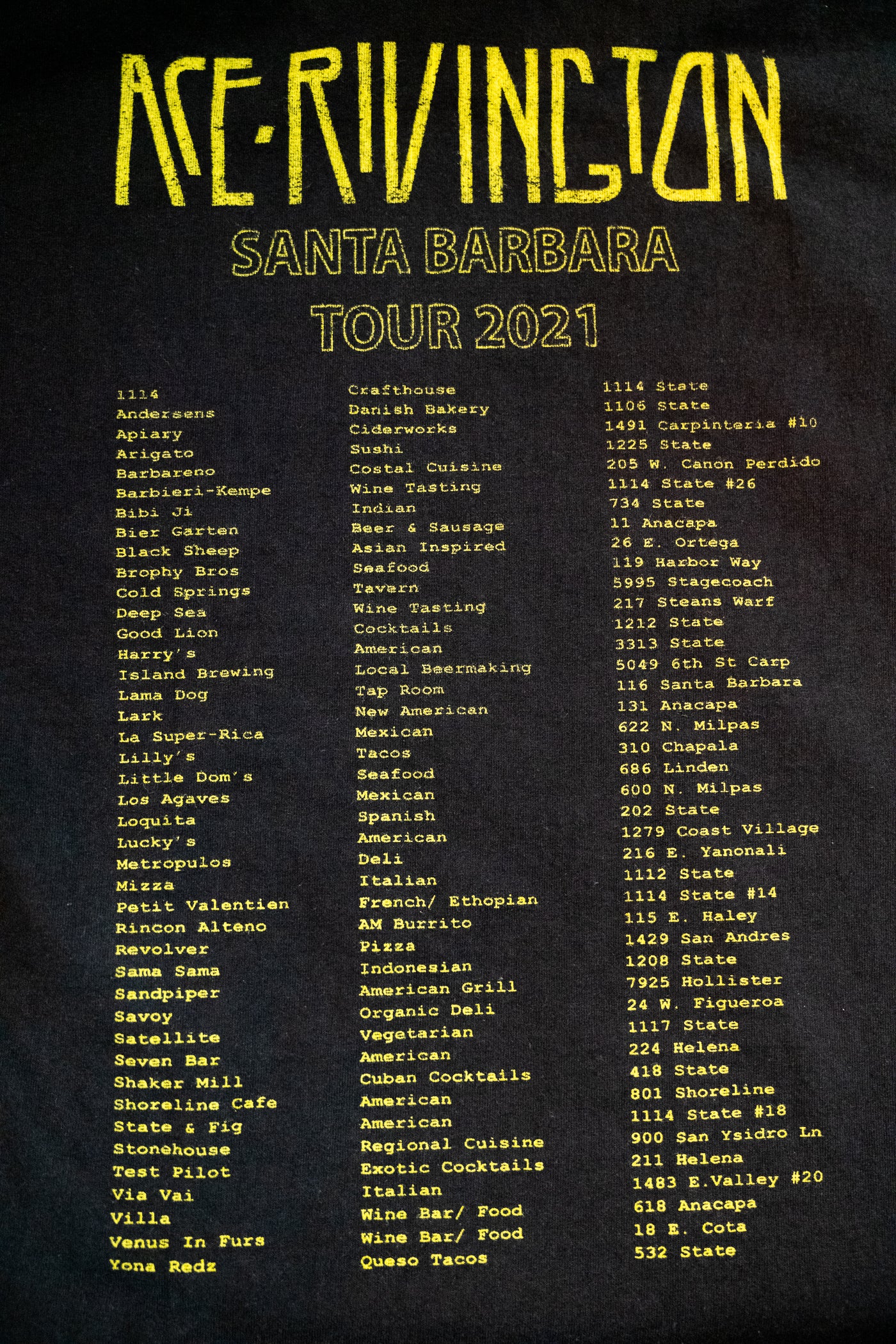 The back of a black color Ace Rivington Heavy Tee - Crewneck SS from the 2021 Monster Tour  t-shirt features a list of various locations in Santa Barbara, organized into three columns under the headings 'Ace Rivington,' 'Santa Barbara,' and 'Tour 2021.' The text is printed in yellow, highlighting the unique tour concept.